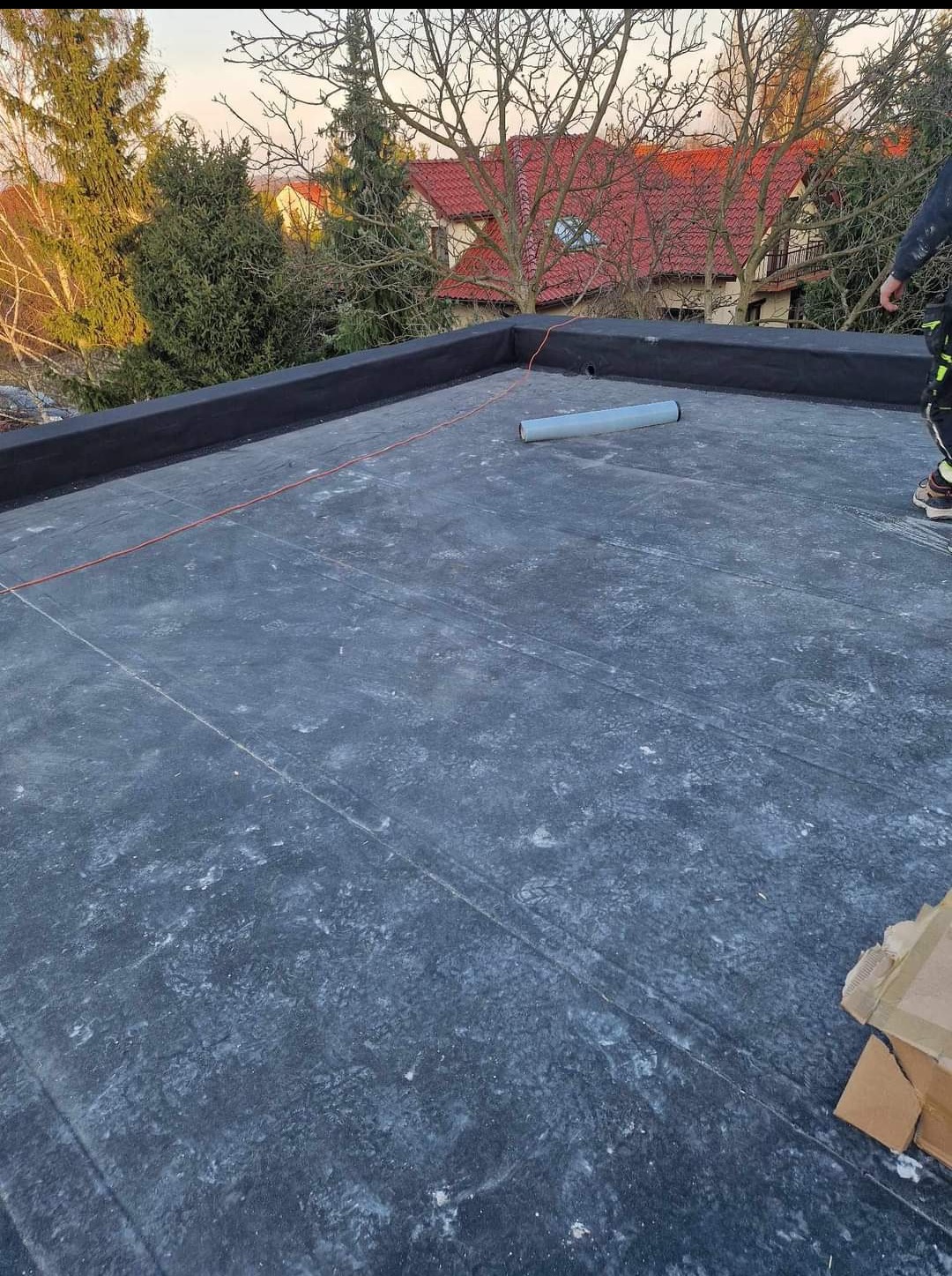 Wykończony płaski dach pokryty czarną papą, widoczny zwój papy, obróbki blacharskie i częściowo postać dekarza w prawym górnym rogu, w tle drzewa i dom z czerwoną dachówką.