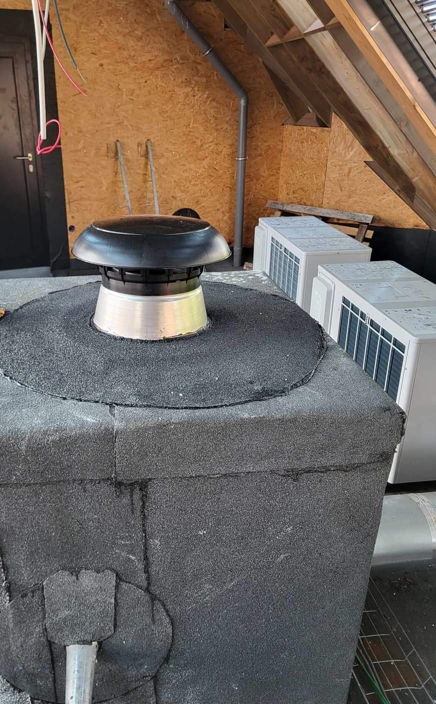 Wykończony komin na dachu pokrytym papą, z zainstalowanym czarnym daszkiem wentylacyjnym i widocznymi jednostkami zewnętrznymi klimatyzacji w tle.