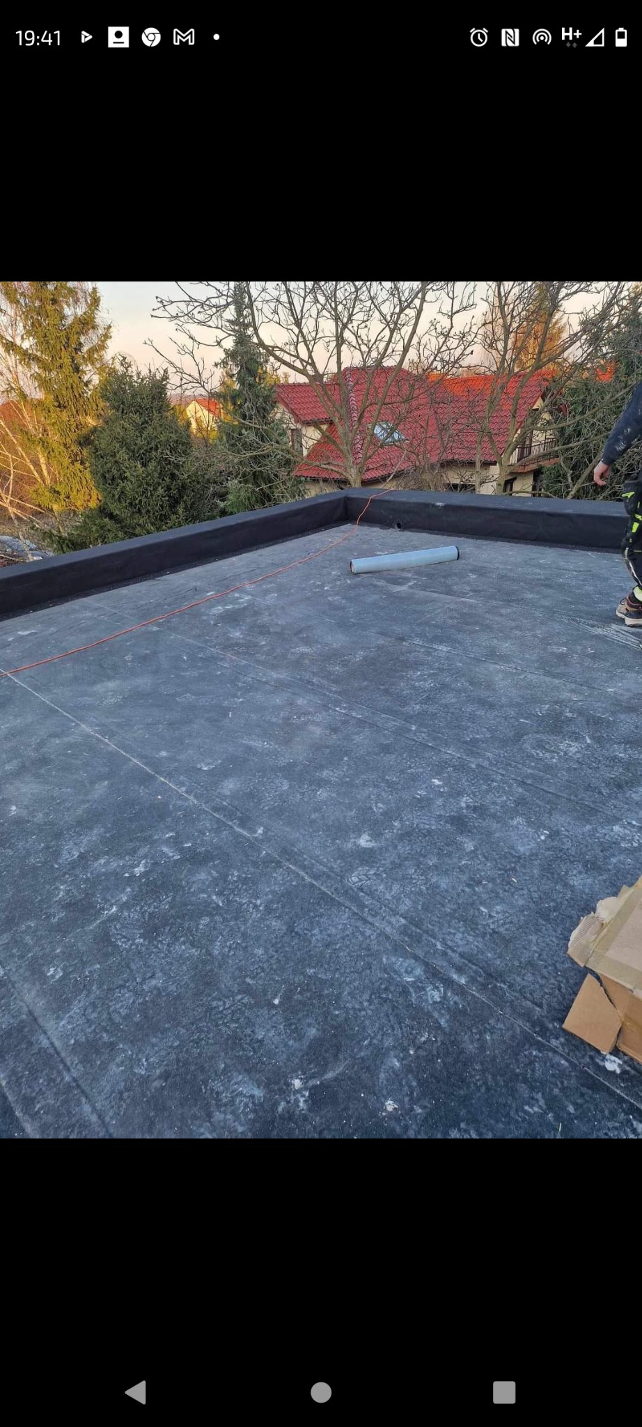Płaski dach pokryty czarną papą, widoczne ślady stóp i rozłożona linka wyznaczająca linię, w tle drzewa i dom z czerwoną dachówką.