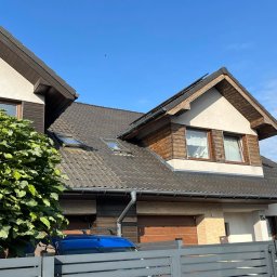 CLEAN HOUSE - Dom jednorodzinny z ciemną dachówką i drewnianą elewacją, widoczny zza metalowego ogrodzenia i bujnego krzewu, z widoczną rynną i fragmentem dachu sąsiedniego budynku z panelami słonecznymi.