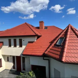 Nowoczesny dom jednorodzinny w Szczecinie z nowym, czerwonym dachem ceramicznym i oknami dachowymi, widziany z perspektywy ogrodu w słoneczny dzień.