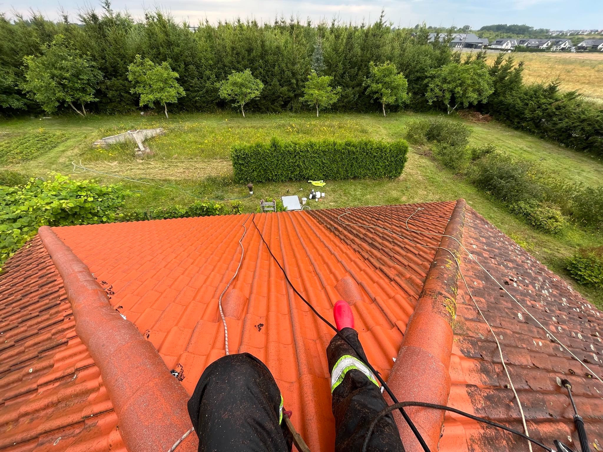 Widok z góry na czerwoną dachówkę podczas czyszczenia, widoczne nogi pracownika w ubraniu roboczym i różowym kaloszu, zieleń drzew w tle.