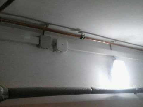 Szara rura biegnąca wzdłuż ściany pod sufitem, z widocznymi puszkami instalacyjnymi i przewodami elektrycznymi, oświetlona intensywnym światłem.