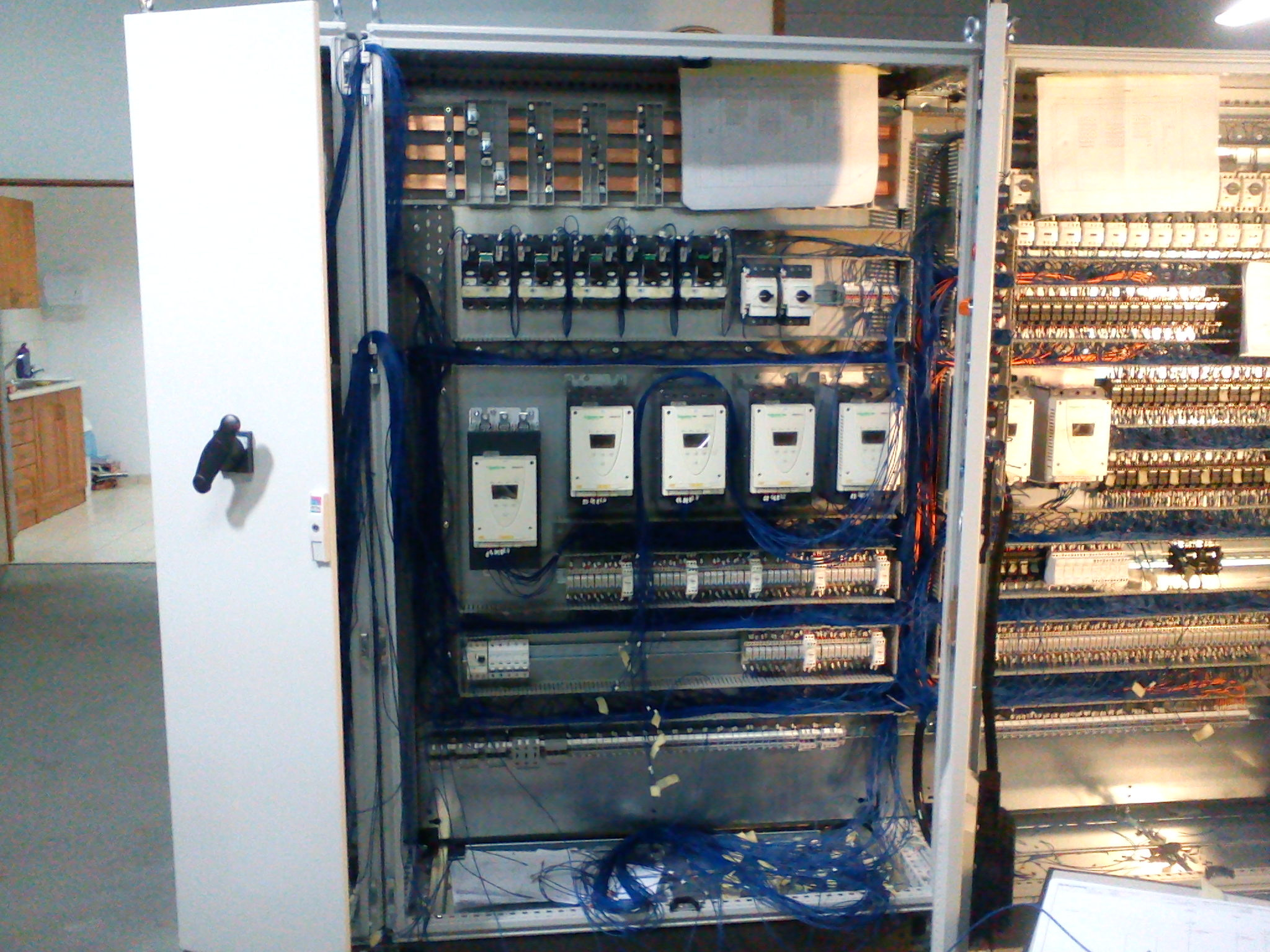 Otwarta szafa elektryczna z widocznymi elementami instalacji, w tym licznymi przewodami, stycznikami i zabezpieczeniami, z uchylonymi drzwiami po lewej stronie.