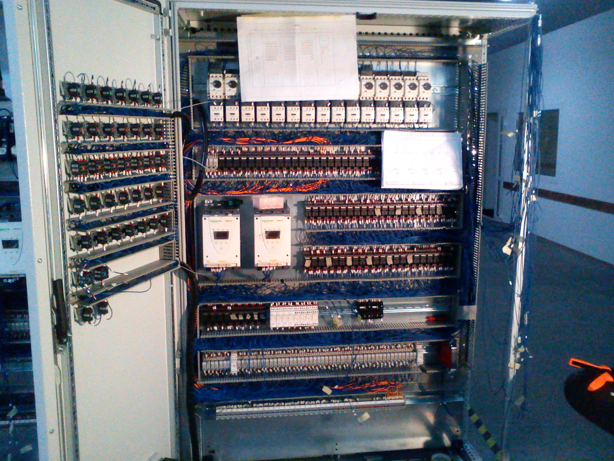 Otwarta szafa sterownicza z licznymi przekaźnikami, zabezpieczeniami i przewodami, widoczne schematy elektryczne wewnątrz, pomarańczowe narzędzie leży obok szafy.