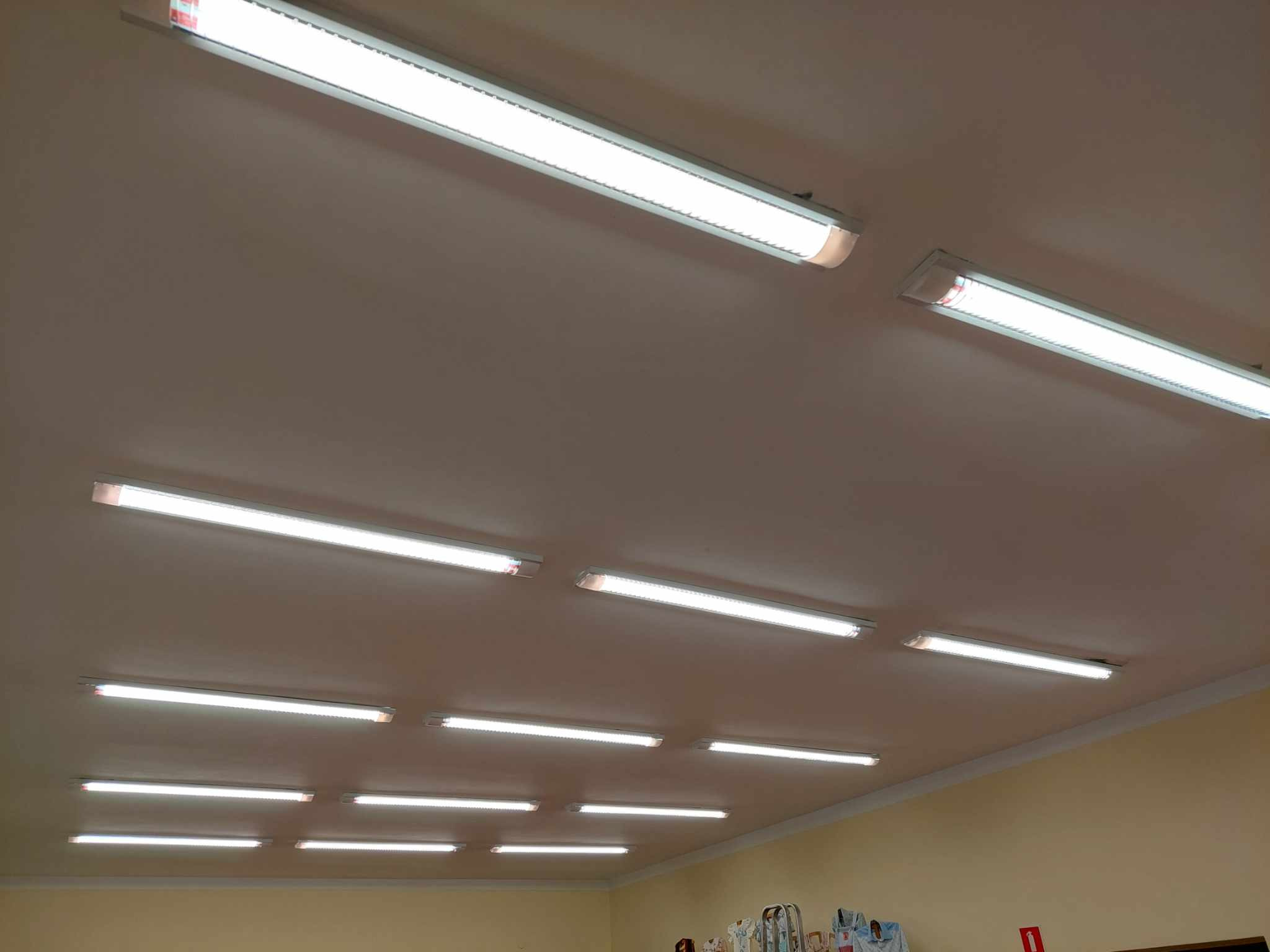 Równomiernie rozmieszczone, podłużne lampy LED zamontowane na suficie w pomieszczeniu o jasnych ścianach, oświetlające przestrzeń biurową lub handlową.