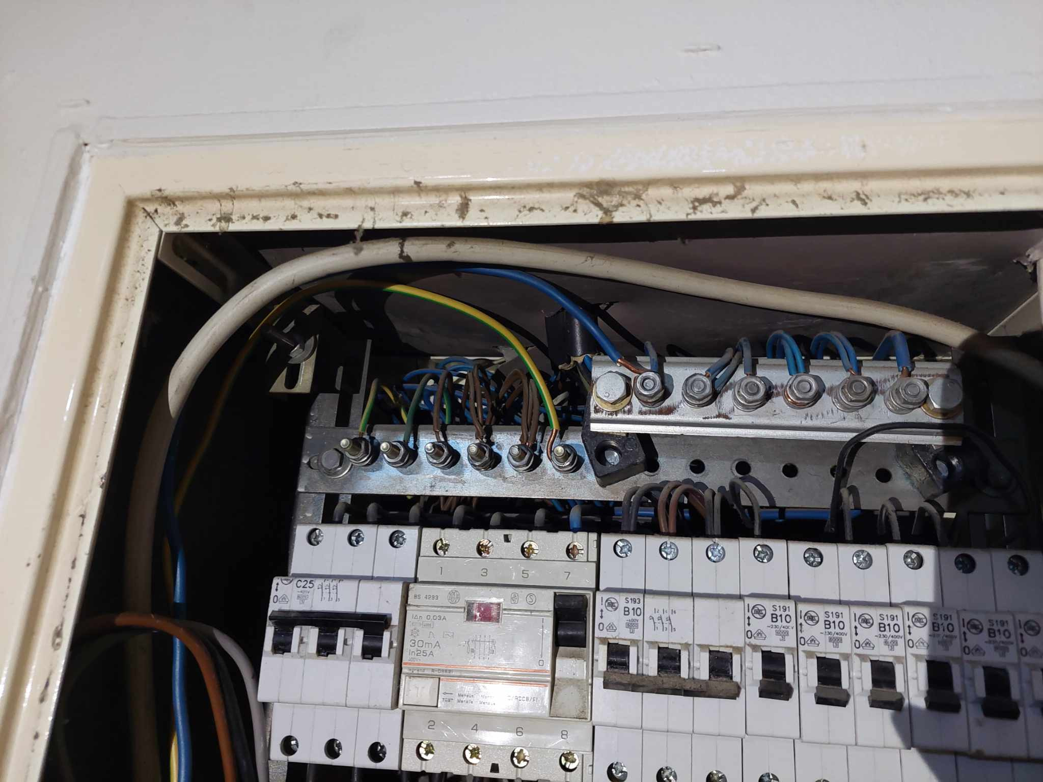 Otwarta skrzynka bezpiecznikowa z widocznymi przewodami, bezpiecznikami i listwami zaciskowymi, z kurzem na obudowie.