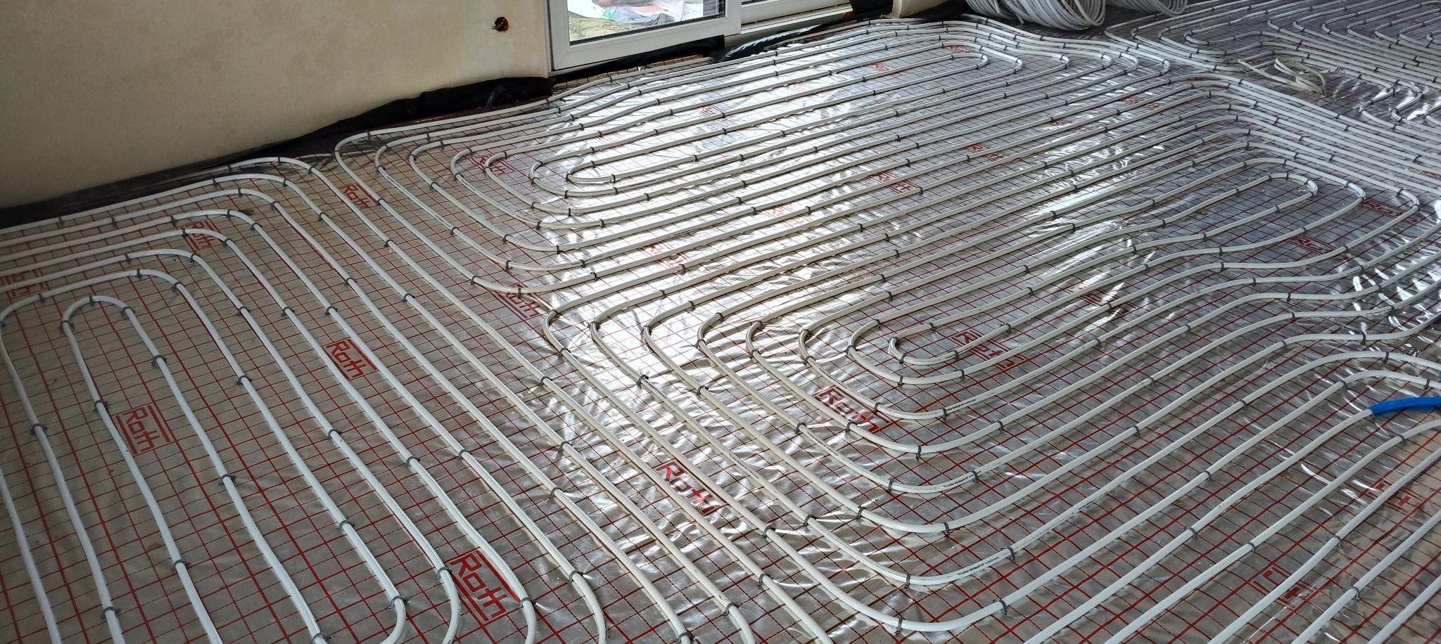 Białe rury ogrzewania podłogowego Roth ułożone na folii z czerwoną siatką, widoczne okno i fragment ściany. Instalacja przed wylaniem posadzki.