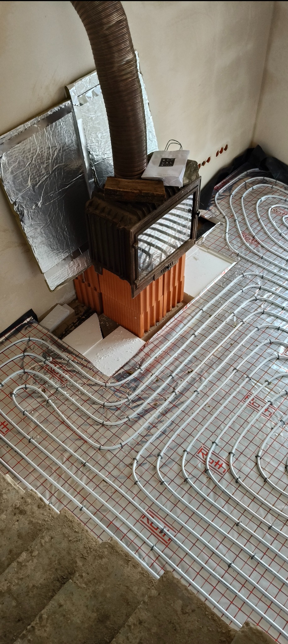 Instalacja ogrzewania podłogowego z widocznym kominem i izolacją, ułożona na folii z siatką. Fragment surowej posadzki w rogu kadru.