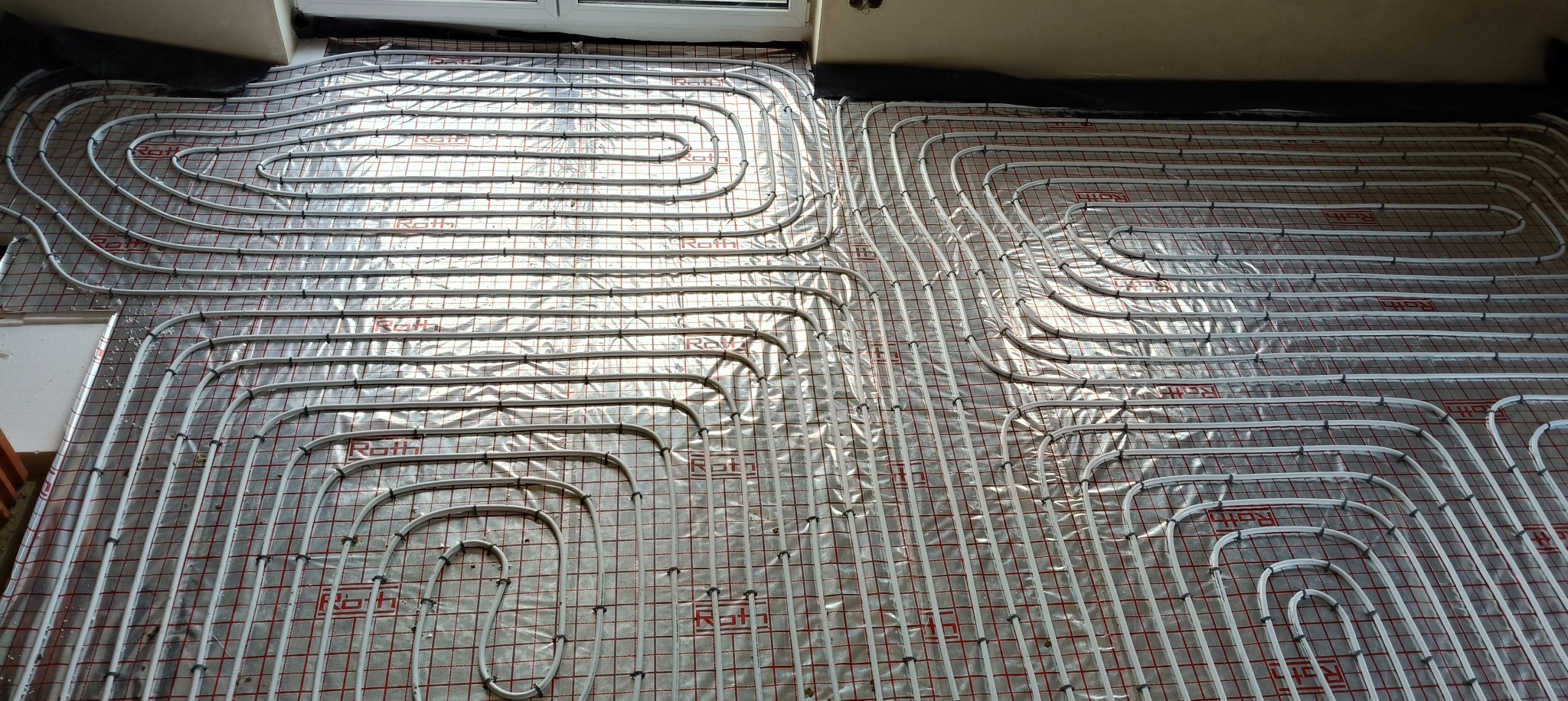 Ułożone rury ogrzewania podłogowego marki Roth na folii z siatką w kolorze czerwonym, widok z góry, etap instalacji przed wylaniem posadzki.