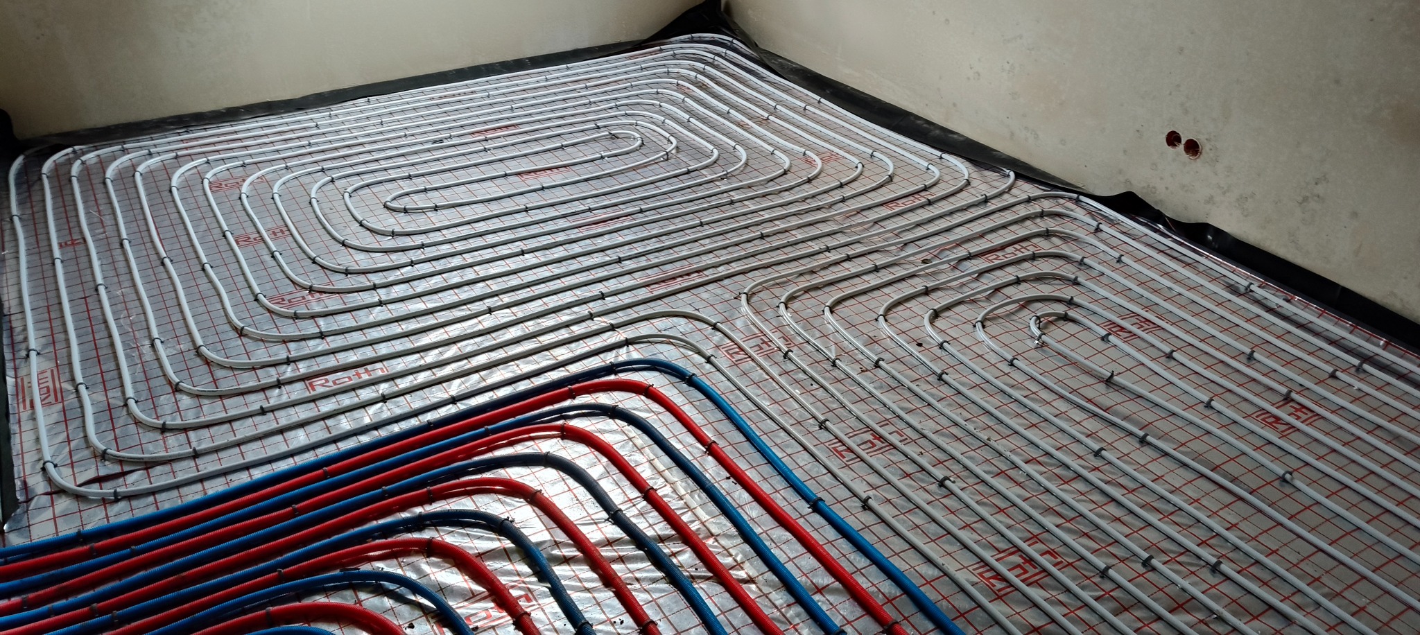Ułożone pętle ogrzewania podłogowego Roth na folii z siatką, z widocznymi rurami zasilającymi (czerwone i niebieskie) w surowym wnętrzu.
