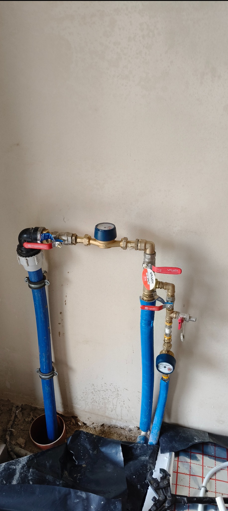 Niebieskie rury instalacji wodnej z zaworami i licznikiem, zamontowane przy ścianie w trakcie prac instalacyjnych. Widoczne narzędzia i folia ochronna na podłodze.