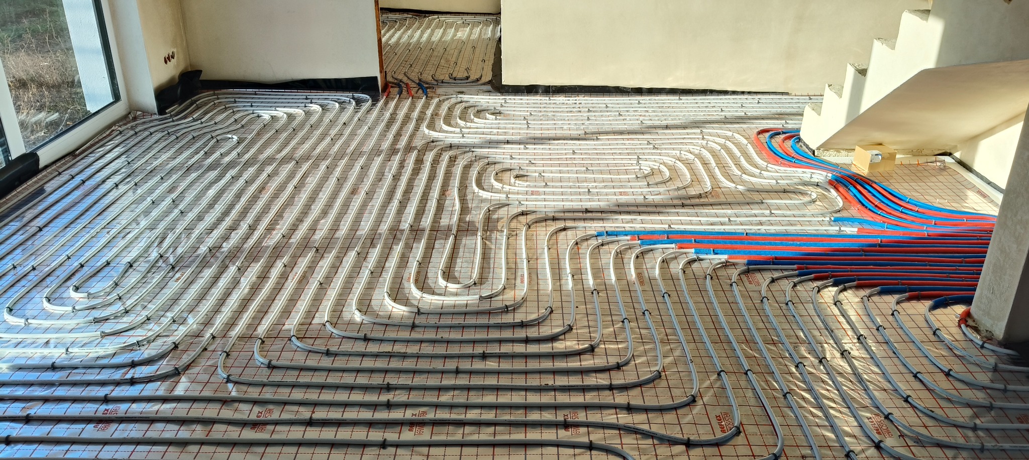Białe rury ogrzewania podłogowego ułożone na folii z siatką, widoczne niebieskie i czerwone rury przy schodach, okno w tle, surowy stan.