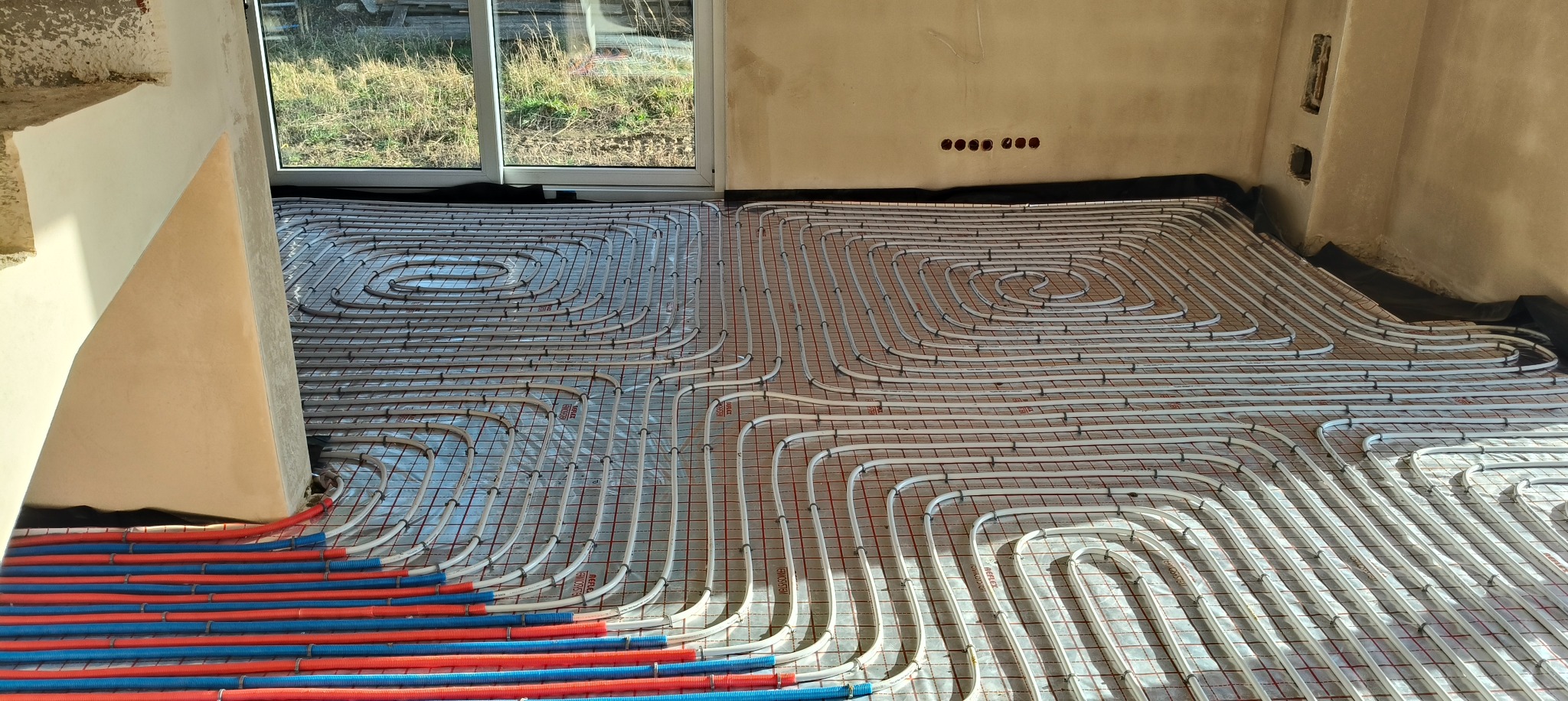 Rozłożona instalacja ogrzewania podłogowego z rurami w kolorze białym, czerwonym i niebieskim na folii izolacyjnej w surowym wnętrzu budynku.