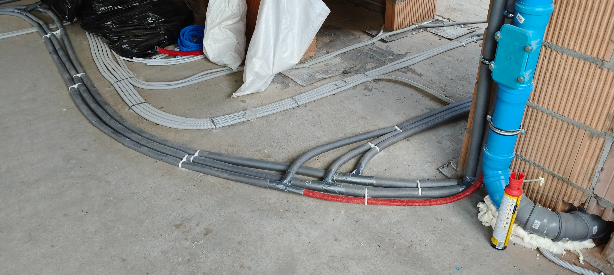 Szare i białe rury instalacyjne ułożone na betonowej podłodze w trakcie montażu, z niebieską rurą pionową przy ścianie z cegły dziurawki. Widoczna puszka pianki montażowej.
