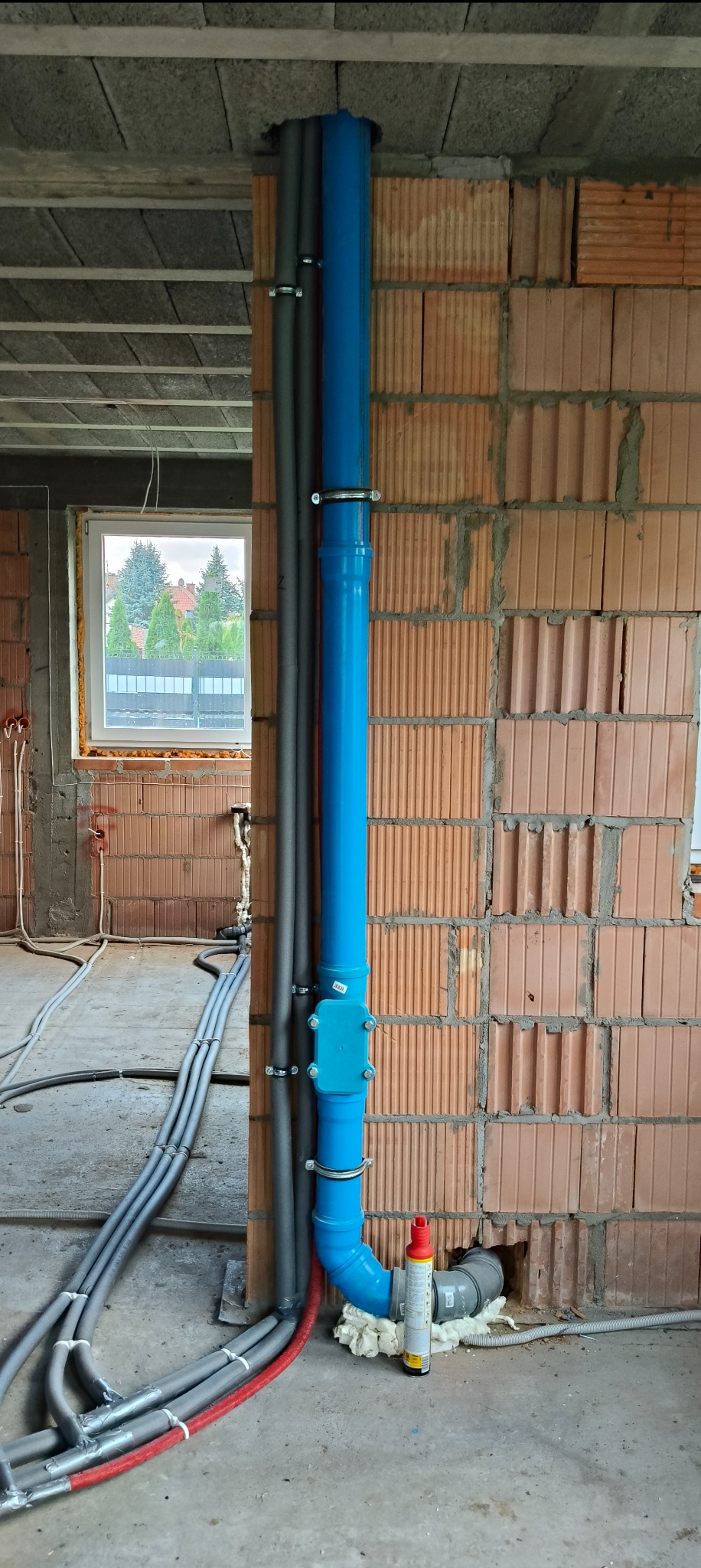 Niebieska rura kanalizacyjna pionowo zamocowana do ściany z cegły dziurawki, szare rury w izolacji, pianka montażowa, widok na okno i budynki w tle.