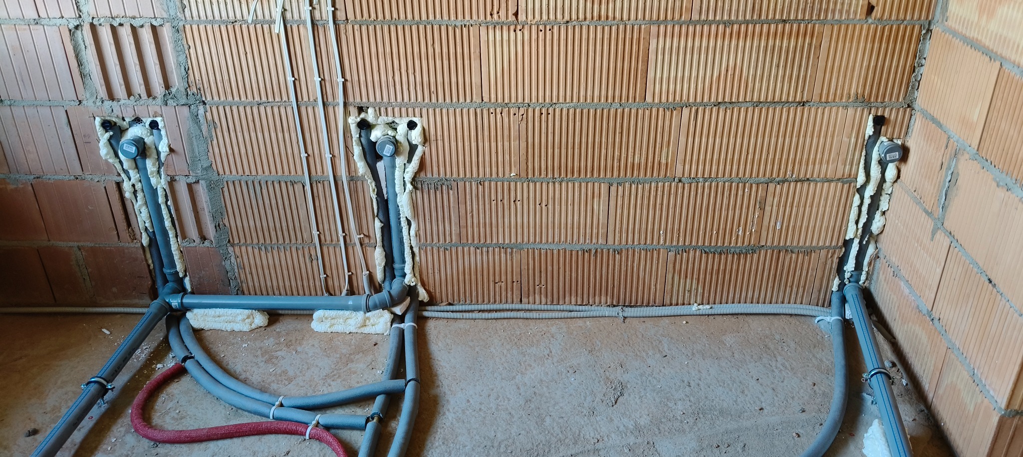 Instalacja wodno-kanalizacyjna w surowym stanie, rury szare i czerwone prowadzone wzdłuż ściany z cegły dziurawki, zabezpieczone pianką montażową.