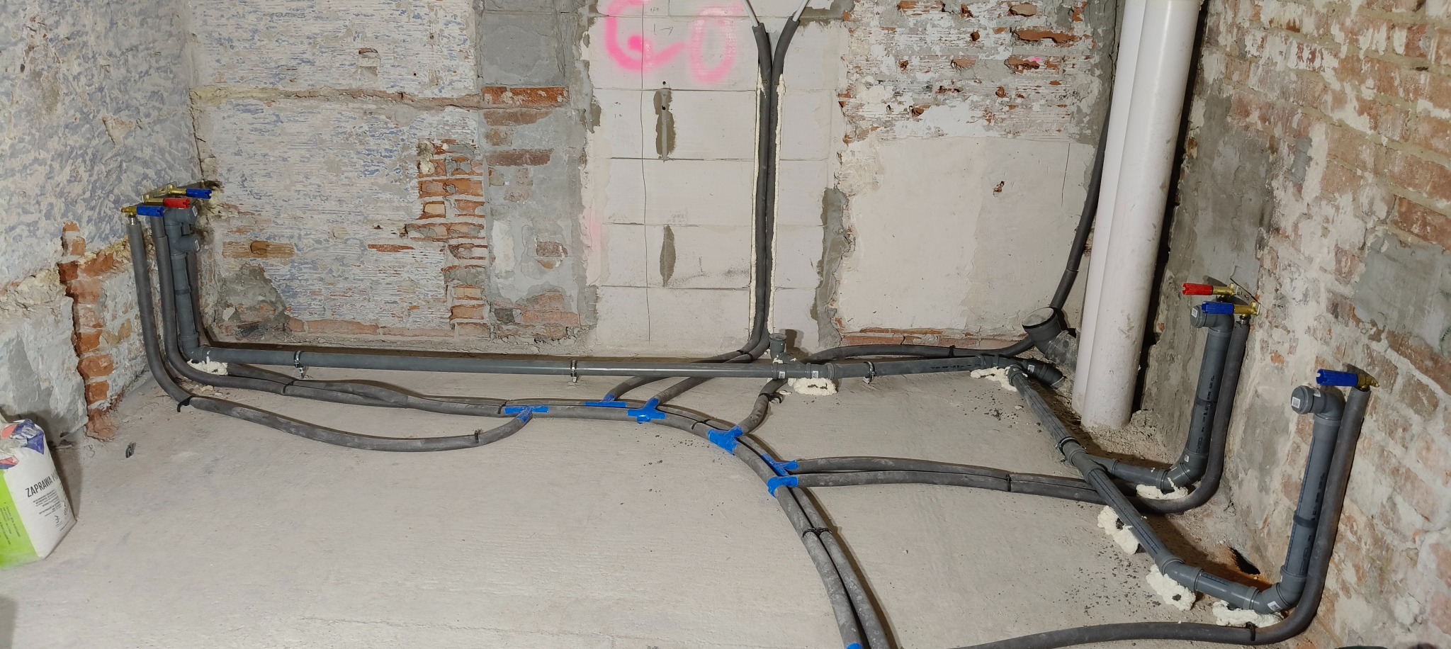 Szare rury instalacji hydraulicznej ułożone na surowym betonie w pomieszczeniu z odsłoniętą cegłą i fragmentami tynku. Widoczne zawory i pianka montażowa.