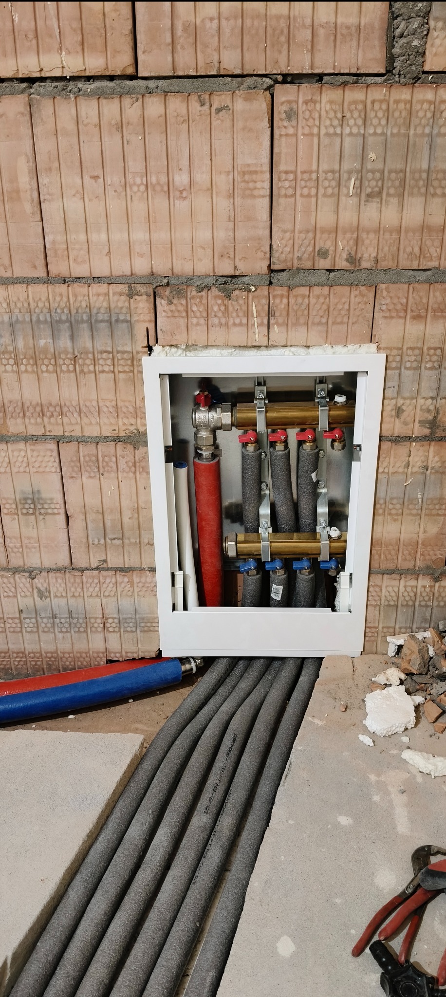 Rozdzielacz ogrzewania podłogowego w białej szafce, widoczne rury w izolacji termicznej, ściana z cegły dziurawki i narzędzia hydrauliczne w tle.