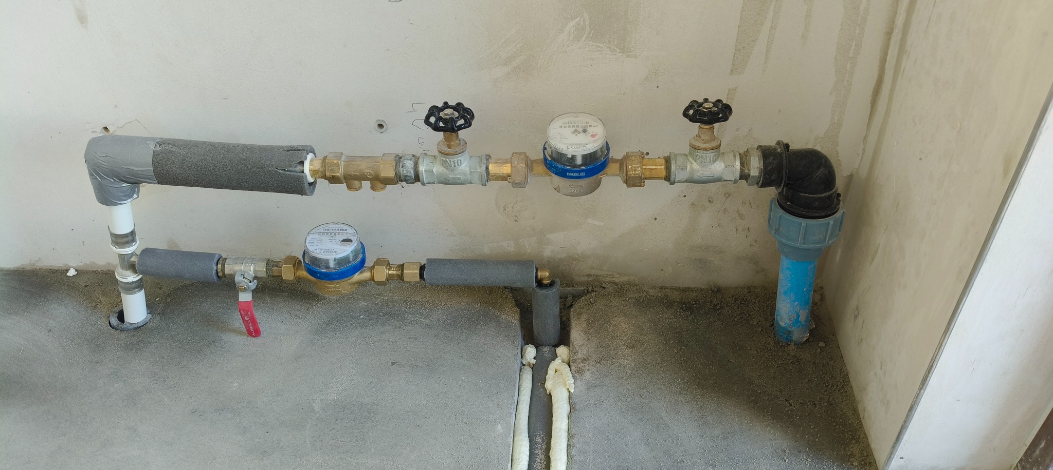 Dwa poziomy instalacji wodnej z licznikami, zaworami i izolacją termiczną na tle surowej ściany i podłogi. Widoczne łączenia i elementy mocujące.