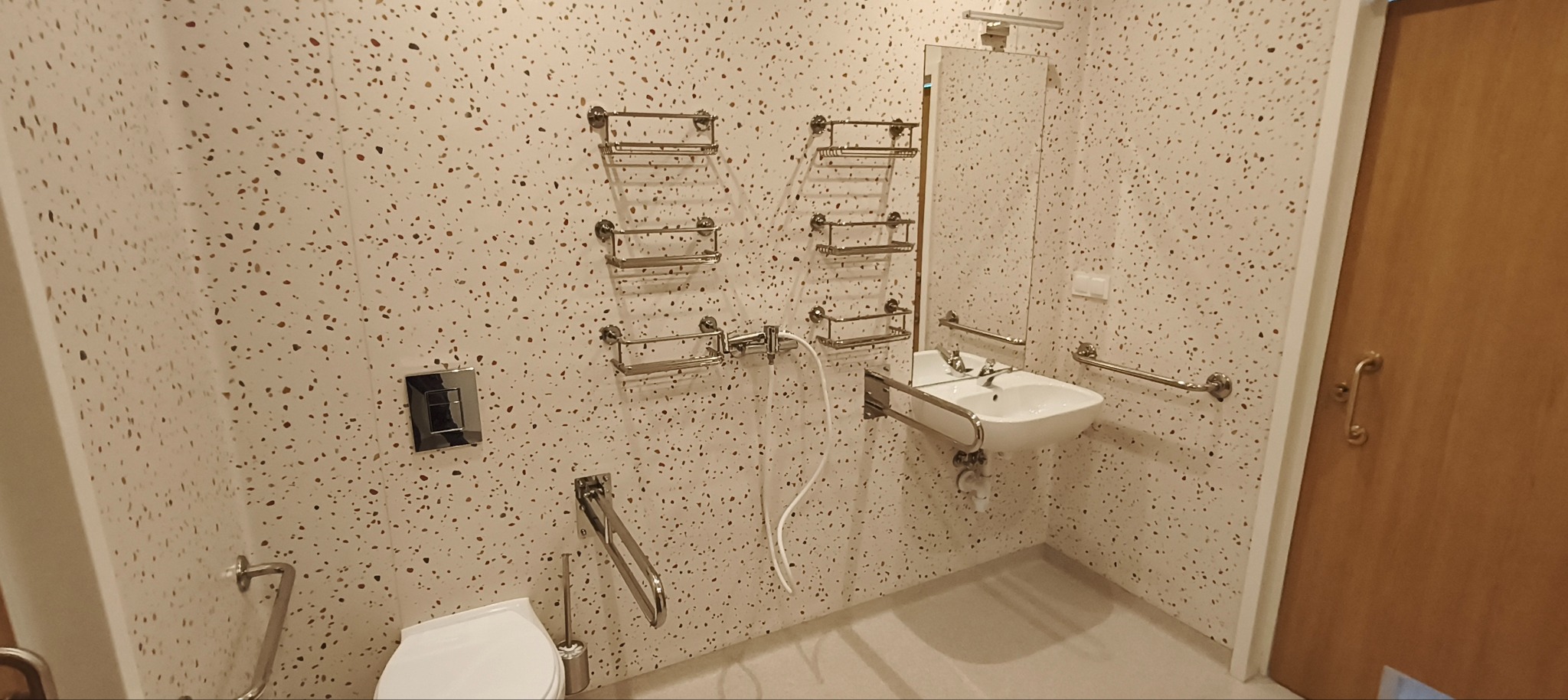 Wykończona łazienka z umywalką i toaletą dla niepełnosprawnych, z chromowanymi uchwytami i półkami na ścianie w kropki. Widoczna armatura i drzwi.