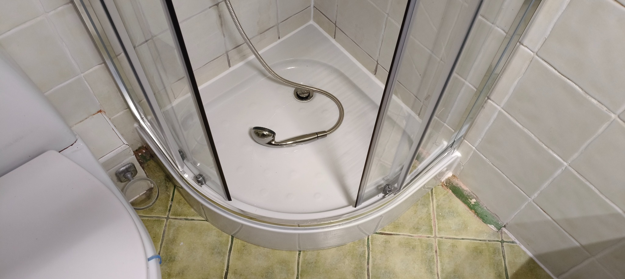 Narożna kabina prysznicowa z chromowaną słuchawką, widok z góry. Biały brodzik i jasne płytki. Częściowo widoczna toaleta po lewej stronie kadru.