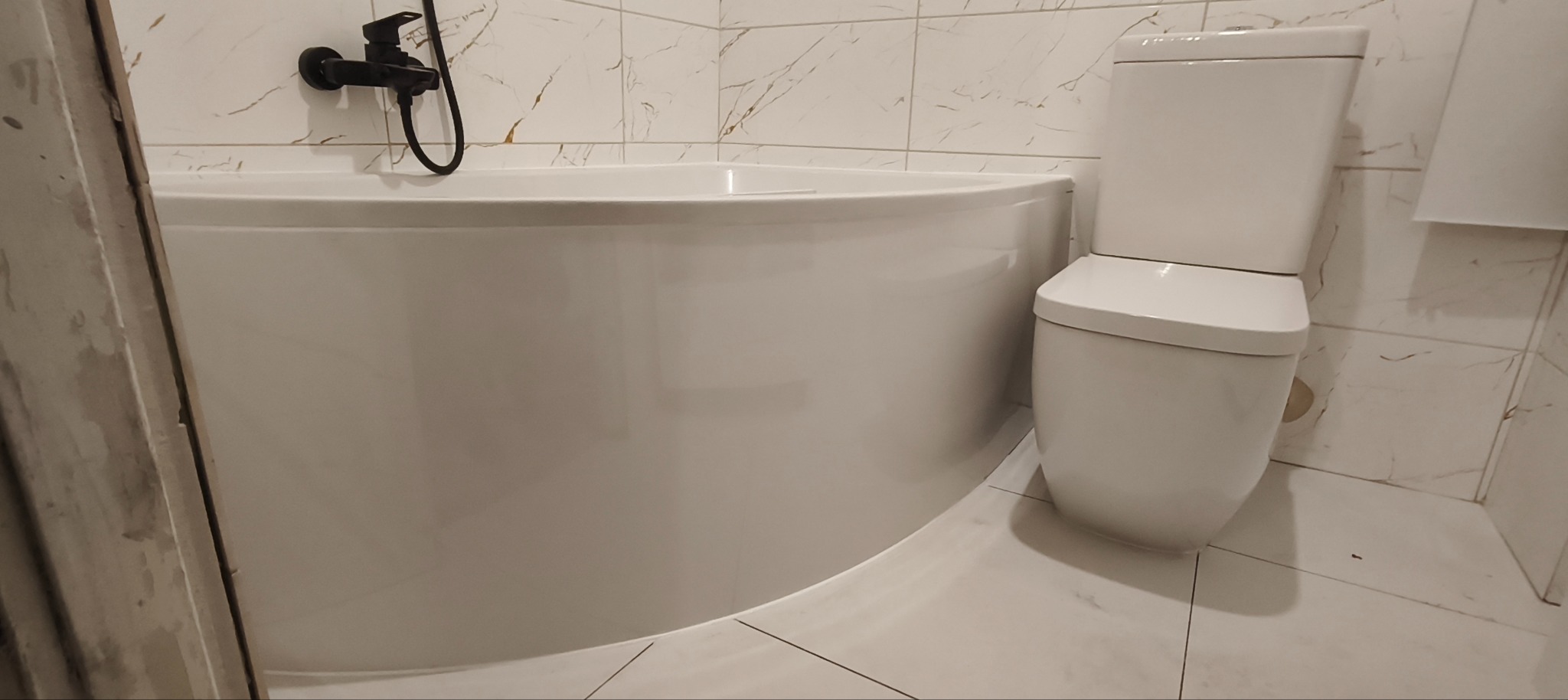 Nowoczesna łazienka z białą, narożną wanną i toaletą. Czarna bateria wannowa na tle marmurowych płytek. Minimalistyczny design, jasne wnętrze.