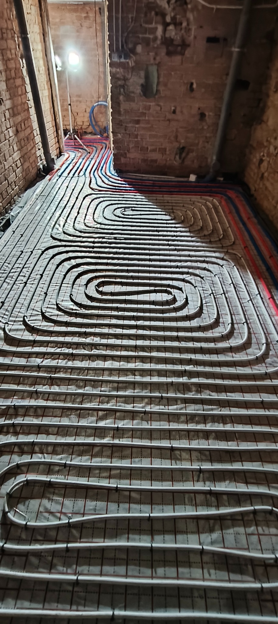 Ułożone pętle ogrzewania podłogowego na izolacji, widoczne rury doprowadzające w kolorze czerwonym i niebieskim. Surowy stan, ceglane ściany w tle.