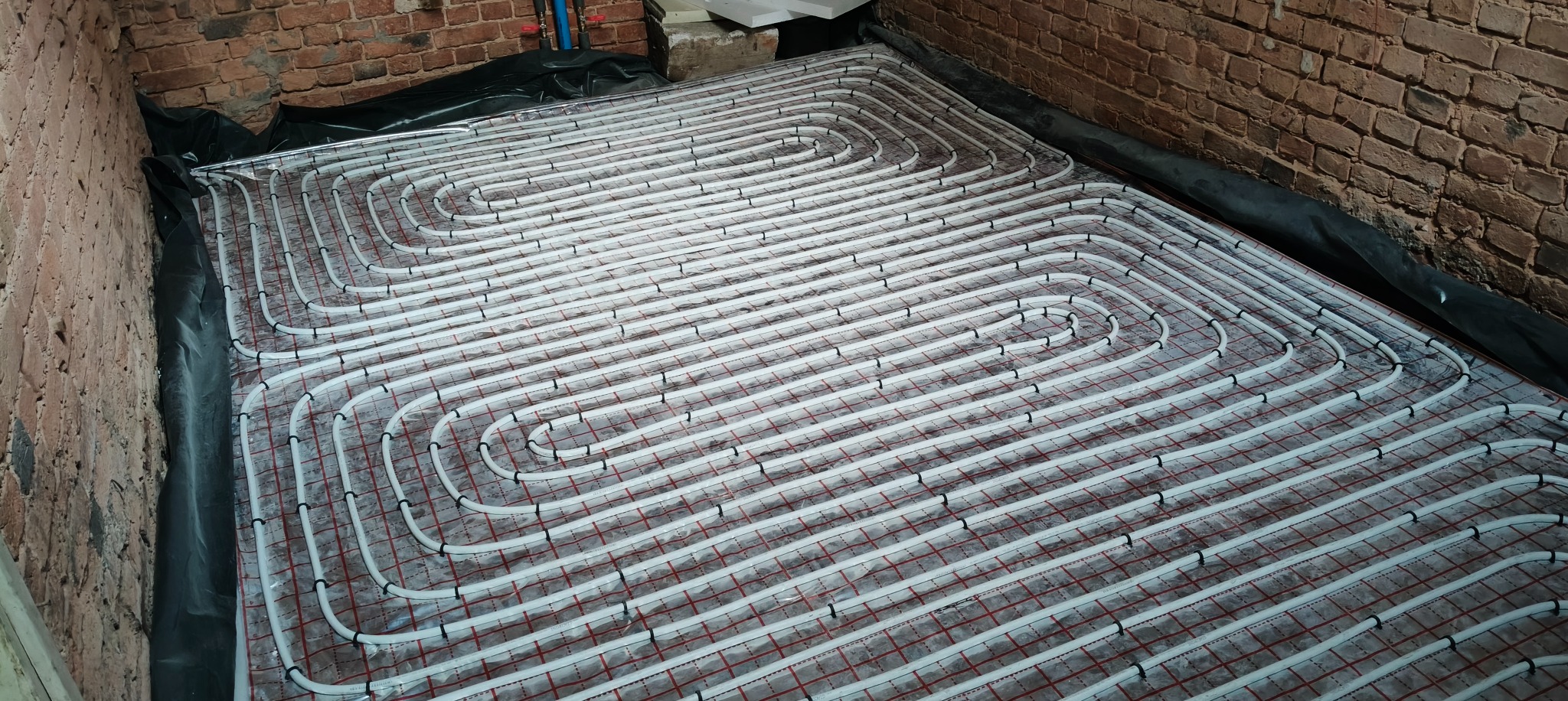 Białe rury ogrzewania podłogowego ułożone spiralnie na czerwonej macie w pomieszczeniu z ceglanymi ścianami i czarną folią izolacyjną. Widok z góry.