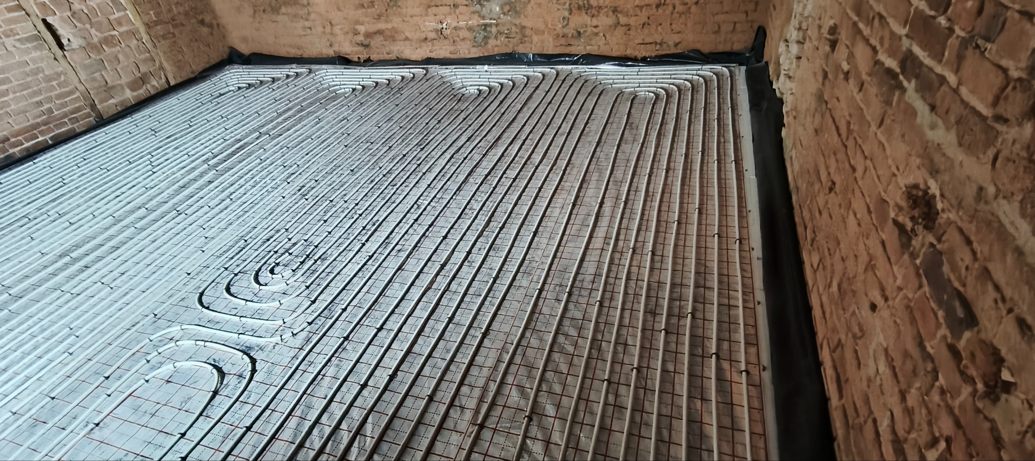 Ułożone białe rury ogrzewania podłogowego na siatce, widoczne ceglane ściany. Instalacja w trakcie realizacji, przed wylaniem posadzki. Ujęcie z góry pod kątem.