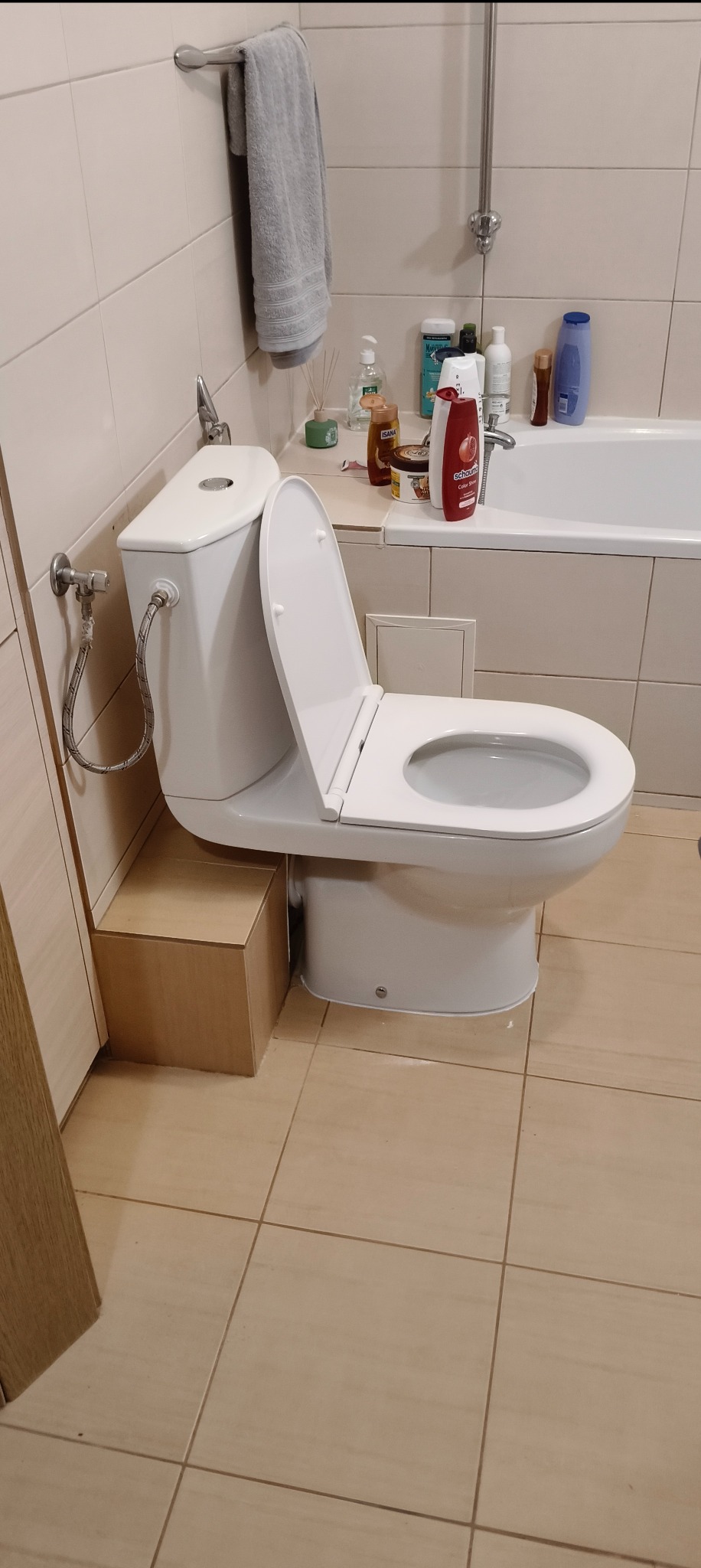 Biała toaleta z otwartą klapą, zamontowana na podwyższeniu w łazience z beżowymi płytkami. Widoczny bidet i akcesoria łazienkowe na wannie.