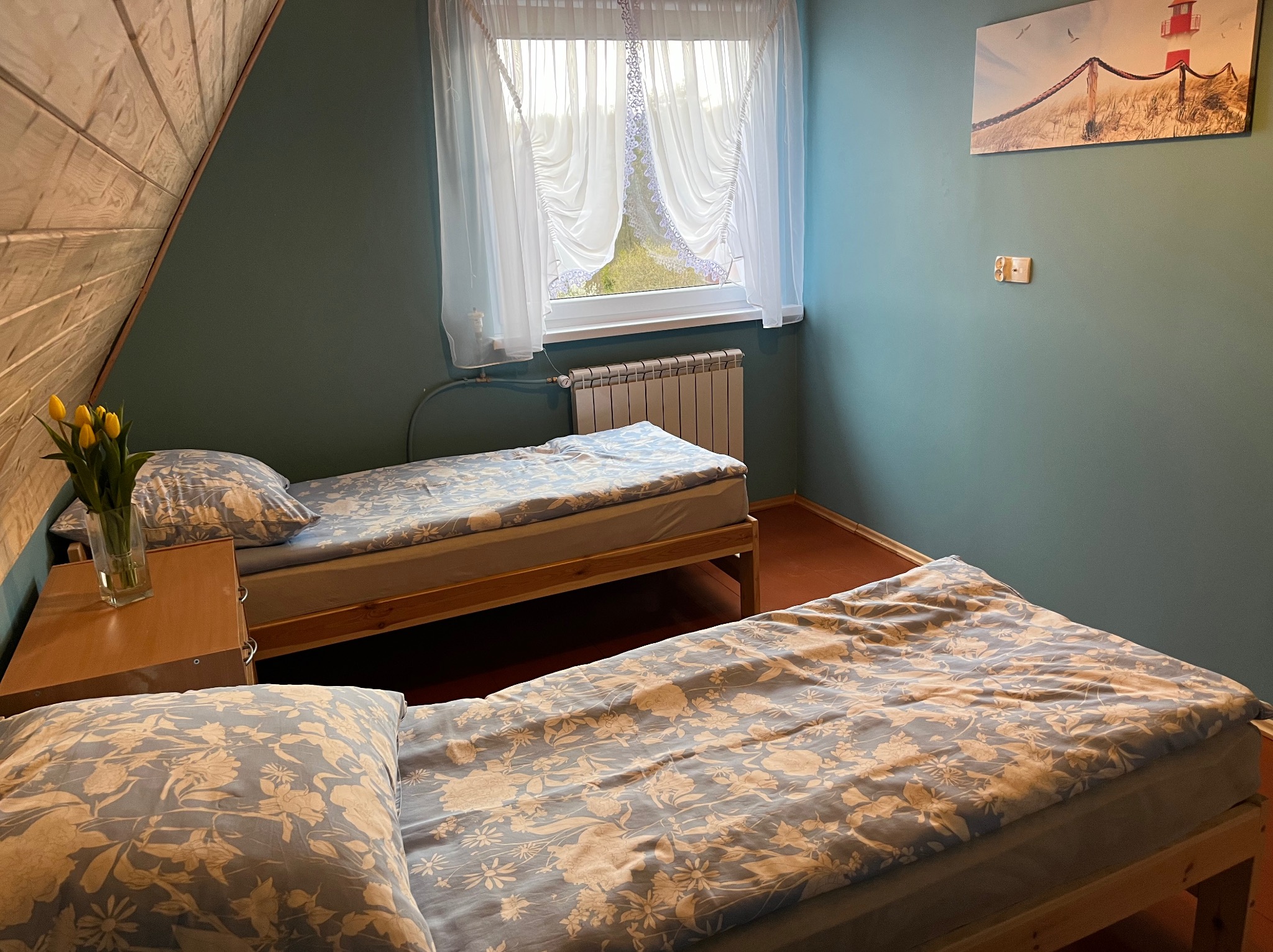 Dwa łóżka pojedyncze z niebieską pościelą w kwiaty w pokoju z niebieskimi ścianami w Kosakowie, widok na okno z firankami i obrazkiem z latarnią morską.