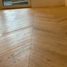 Jasne, drewniane panele ułożone w jodełkę, widoczne przy białej ścianie i oknie balkonowym.