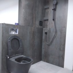 VAD_BUD - Nowoczesna łazienka z wiszącą, czarną toaletą i prysznicem typu deszczownica na ścianie wyłożonej płytkami imitującymi drewno.