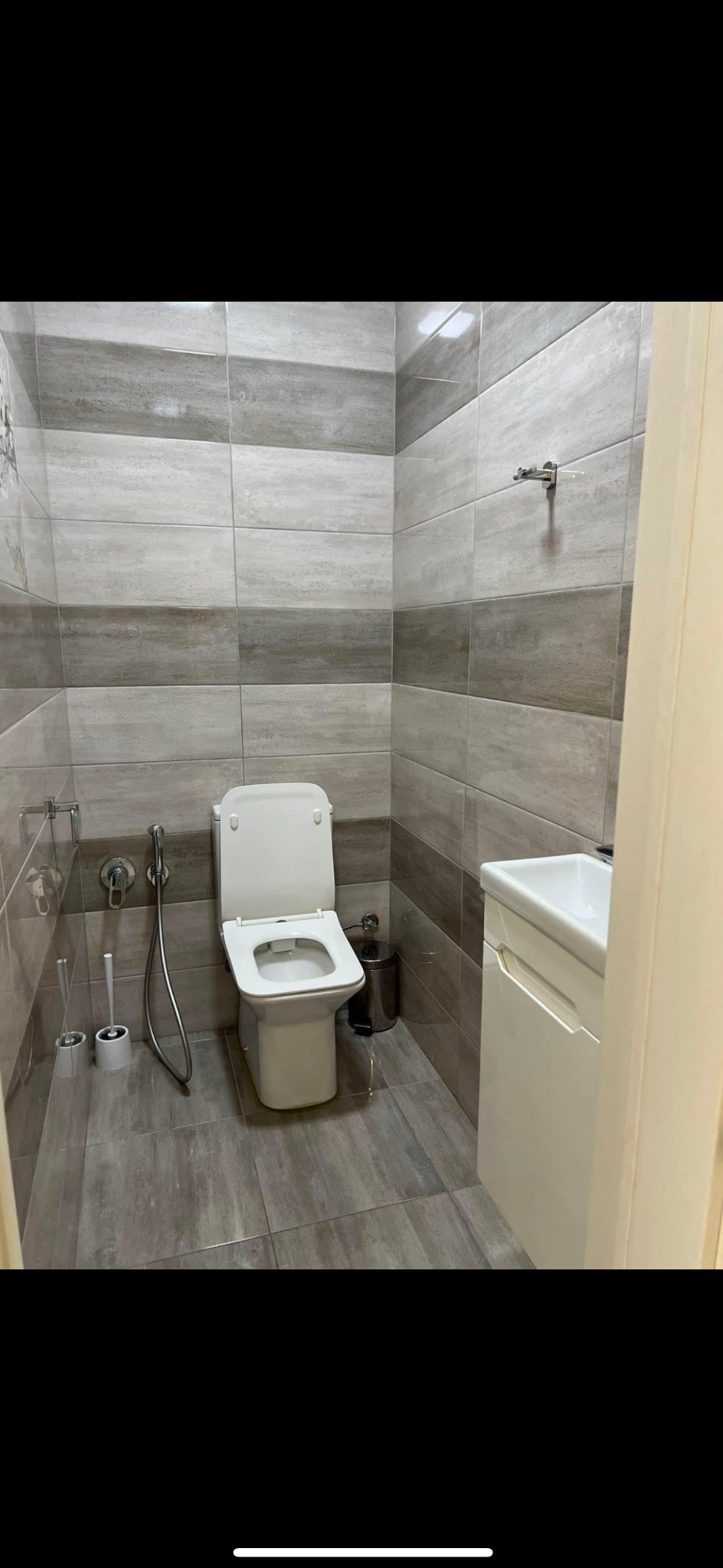 Wnętrze łazienki z toaletą z otwartą klapą, umywalką z szafką i szarymi płytkami imitującymi drewno na ścianach i podłodze.