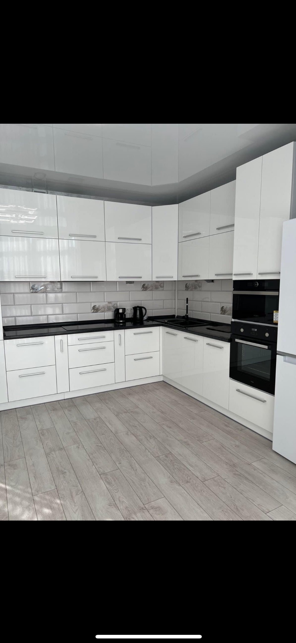 Nowoczesna kuchnia z białymi, lakierowanymi szafkami, czarnym blatem i szarą, cegiełkową mozaiką nad blatem, uzupełniona drewnopodobną podłogą i czarnymi sprzętami.