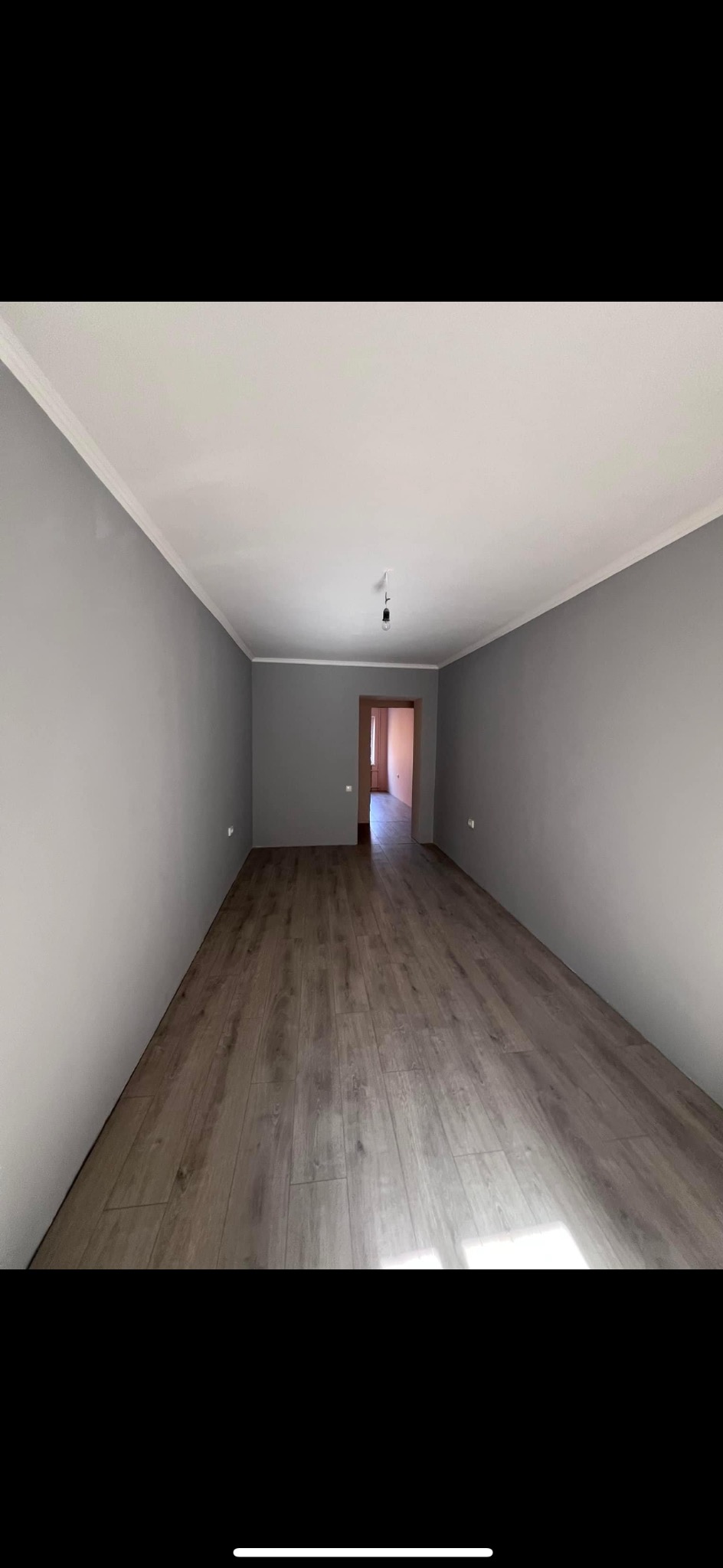 Pomalowane na szaro wnętrze z ułożonymi panelami podłogowymi, widoczne listwy przypodłogowe i sufitowe, surowa żarówka zwisająca z sufitu.