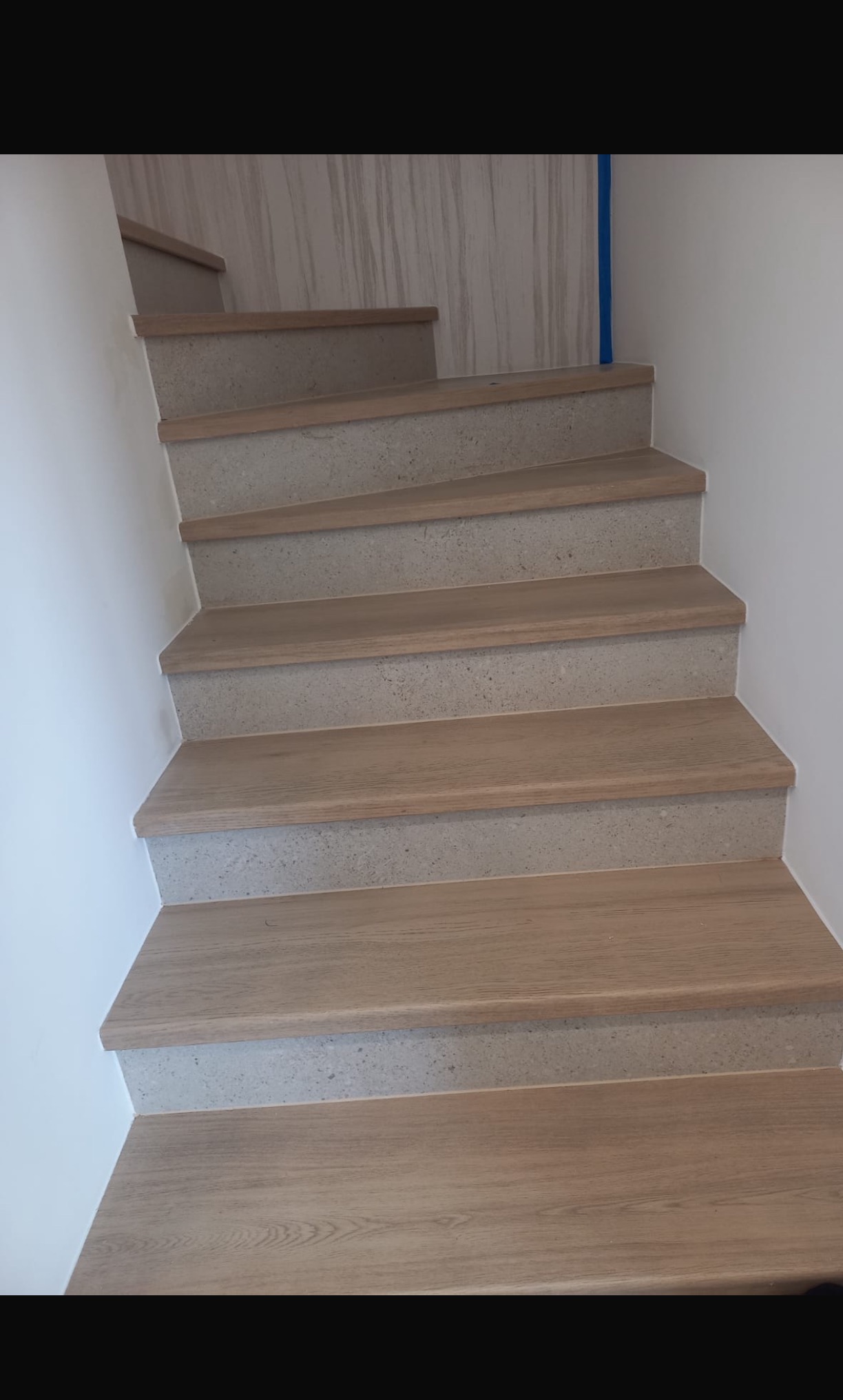 Wykończone schody wewnętrzne z drewnianymi stopniami i betonowymi podstopnicami, widoczne z dołu, z fragmentem marmurowej ściany w tle.