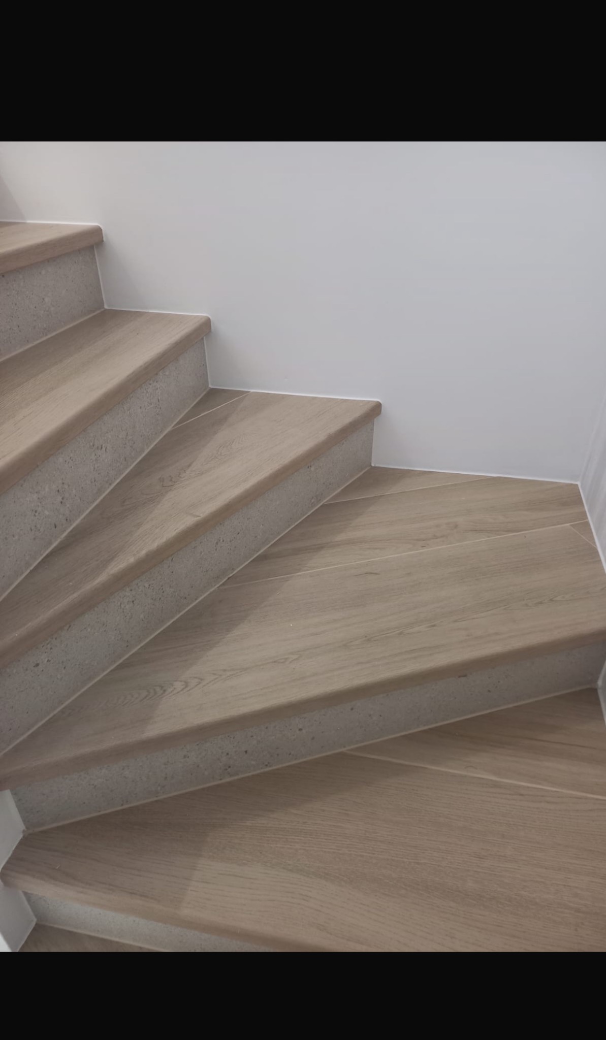 Schody wyłożone płytkami imitującymi jasne drewno, z szarymi, betonowymi krawędziami, prowadzące w górę wzdłuż białej ściany.