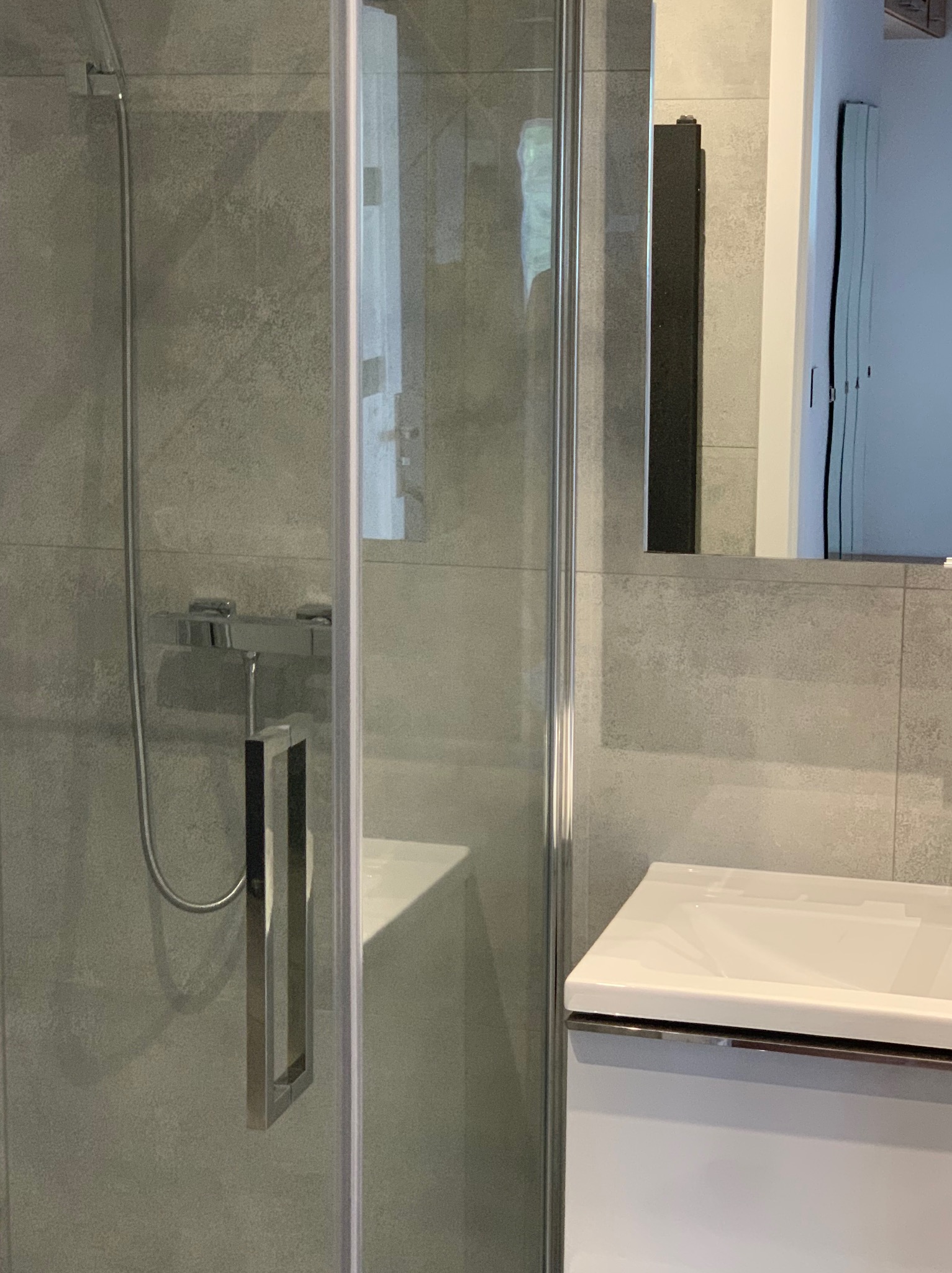 Szklana kabina prysznicowa z chromowanym uchwytem, szara ściana z płytek, biała umywalka z szafką w nowoczesnej łazience.