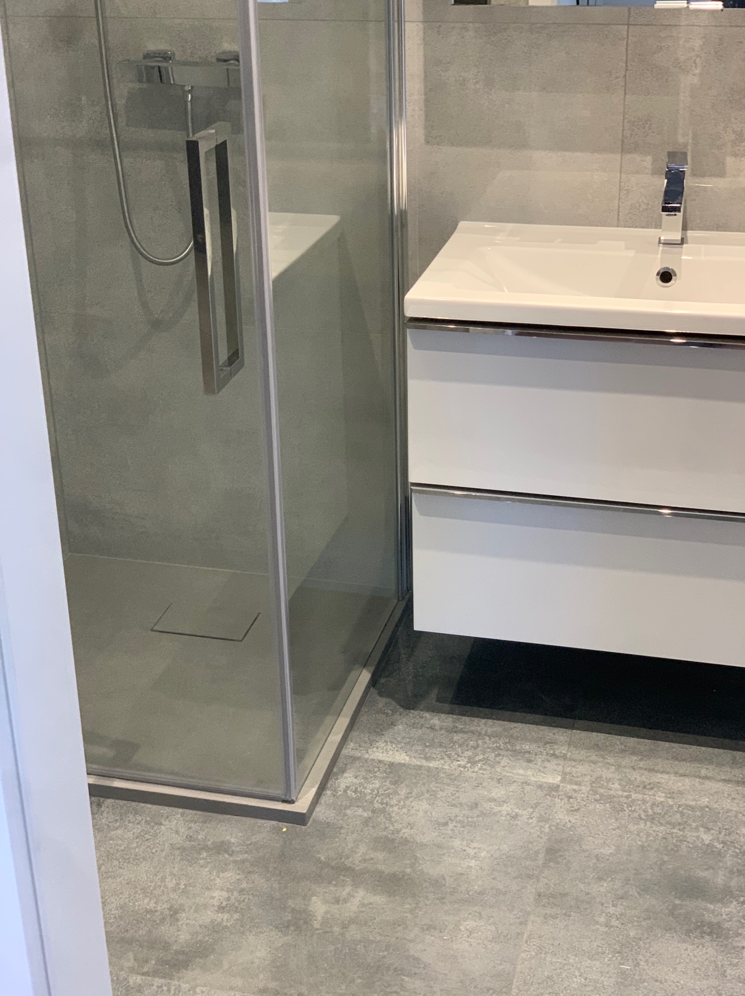 Nowoczesna łazienka z kabiną prysznicową typu walk-in, białą umywalką z szafką i szarą płytką podłogową imitującą beton.