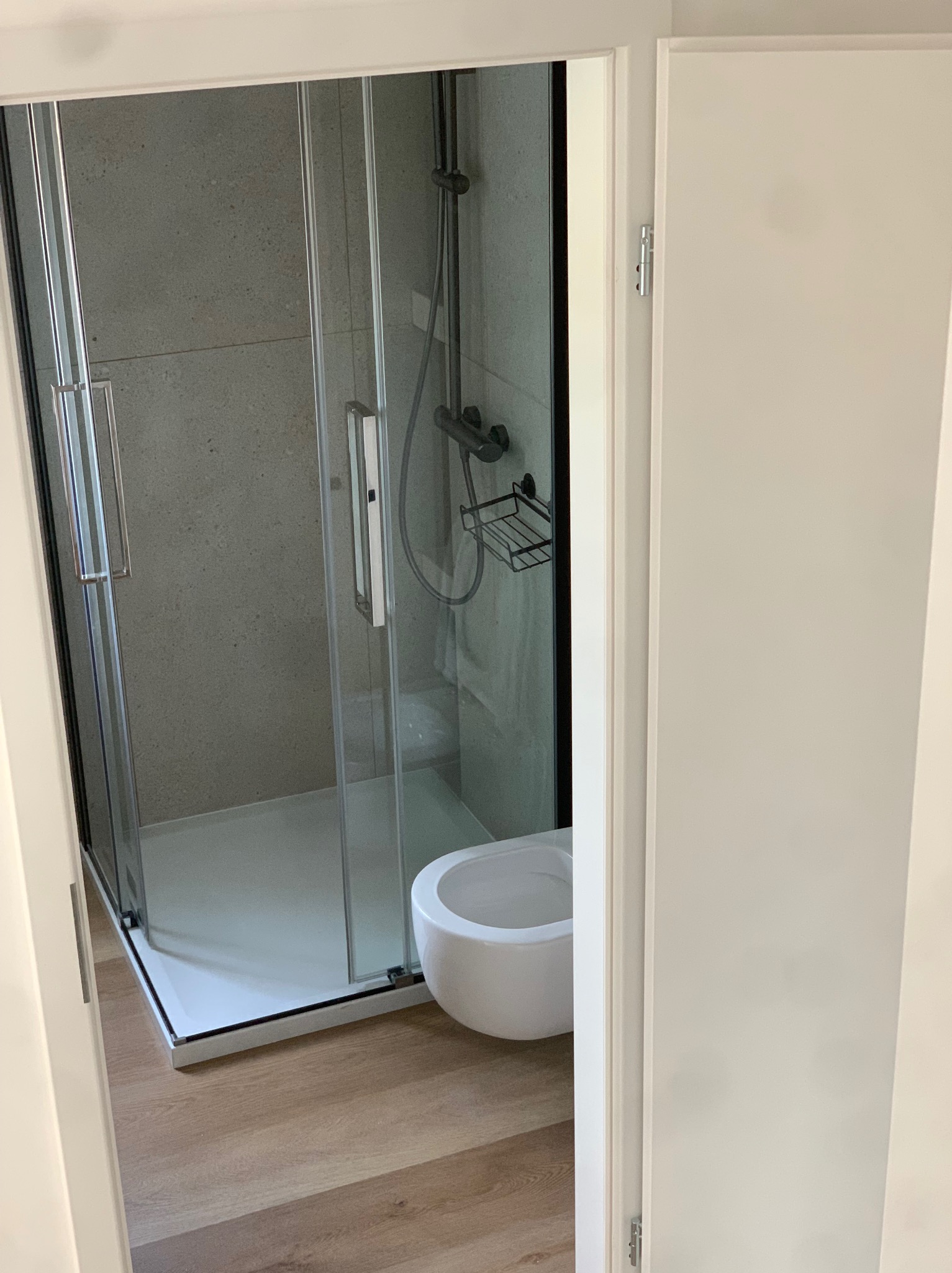 Nowoczesna kabina prysznicowa z czarną armaturą i białą miską WC, widoczna przez otwarte drzwi do łazienki, podłoga wyłożona panelami imitującymi drewno.