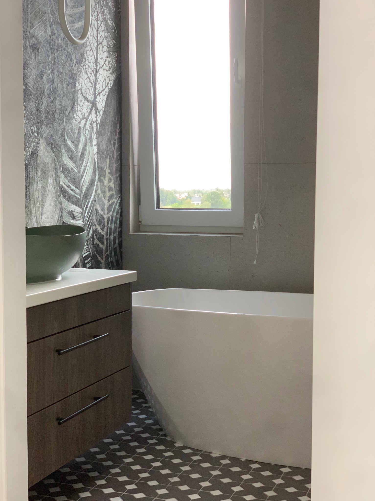 Nowoczesna łazienka z wolnostojącą wanną, umywalką nablatową w kształcie misy, szafką z szufladami w kolorze ciemnego drewna i tapetą w motywy roślinne.