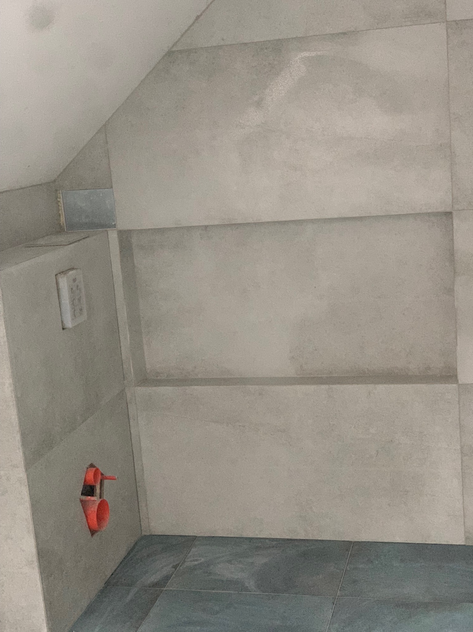 Ściana łazienki z szarymi płytkami w trakcie montażu, widoczne niedokończone przyłącze wodne w kolorze pomarańczowym i wystająca puszka elektryczna.