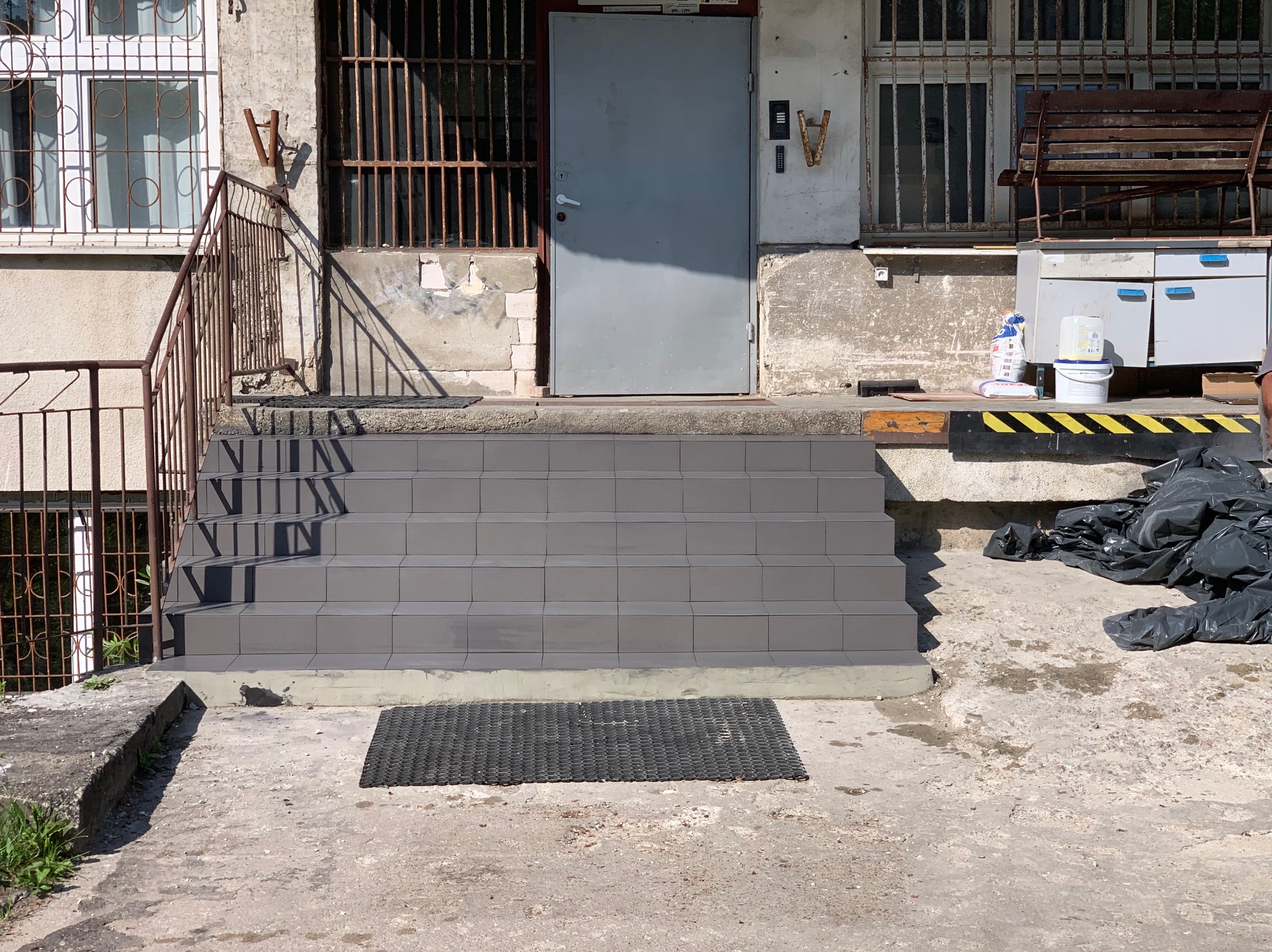 Schody wejściowe wyłożone szarą płytką ceramiczną, z widoczną wycieraczką i fragmentem elewacji budynku z metalowymi oknami.