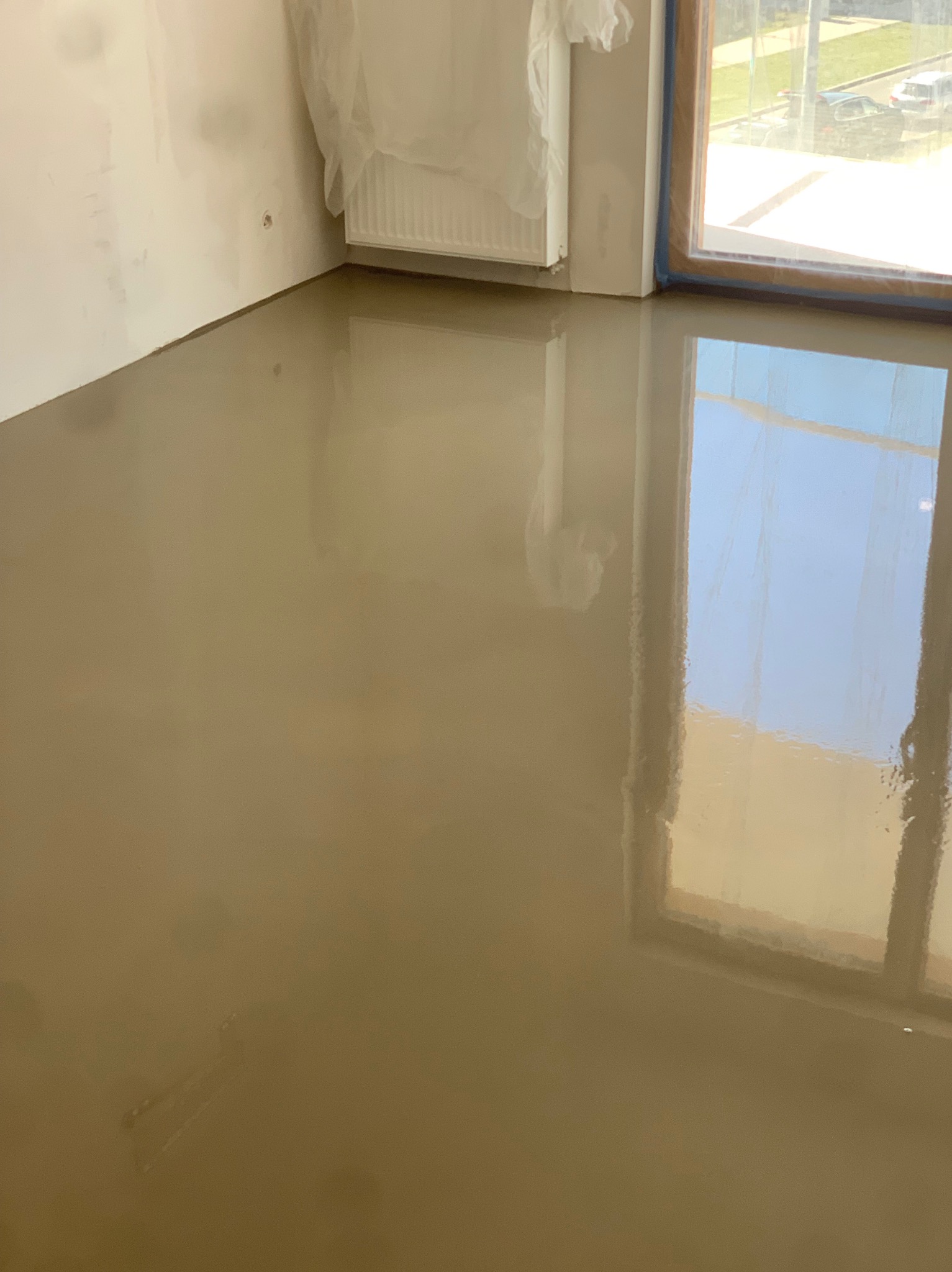 Samopoziomująca wylewka podłogowa w jasnym odcieniu brązu, świeżo wylana, z widocznym odbiciem okna, ściany zabezpieczone folią.