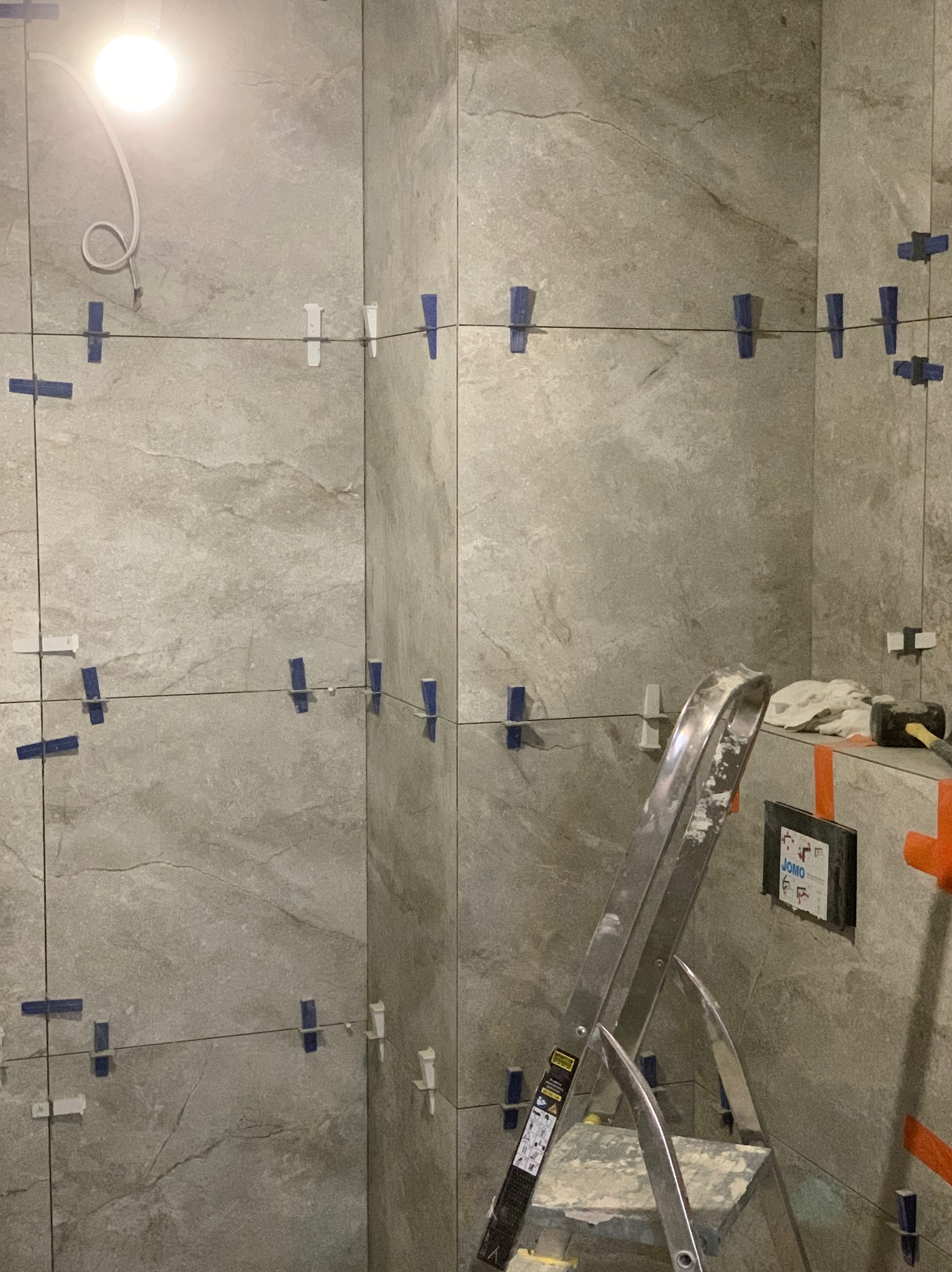 Ściana wyłożona szarymi płytkami imitującymi kamień, z użyciem niebieskich i białych krzyżyków dystansowych, widoczna drabina aluminiowa i narzędzia glazurnicze.