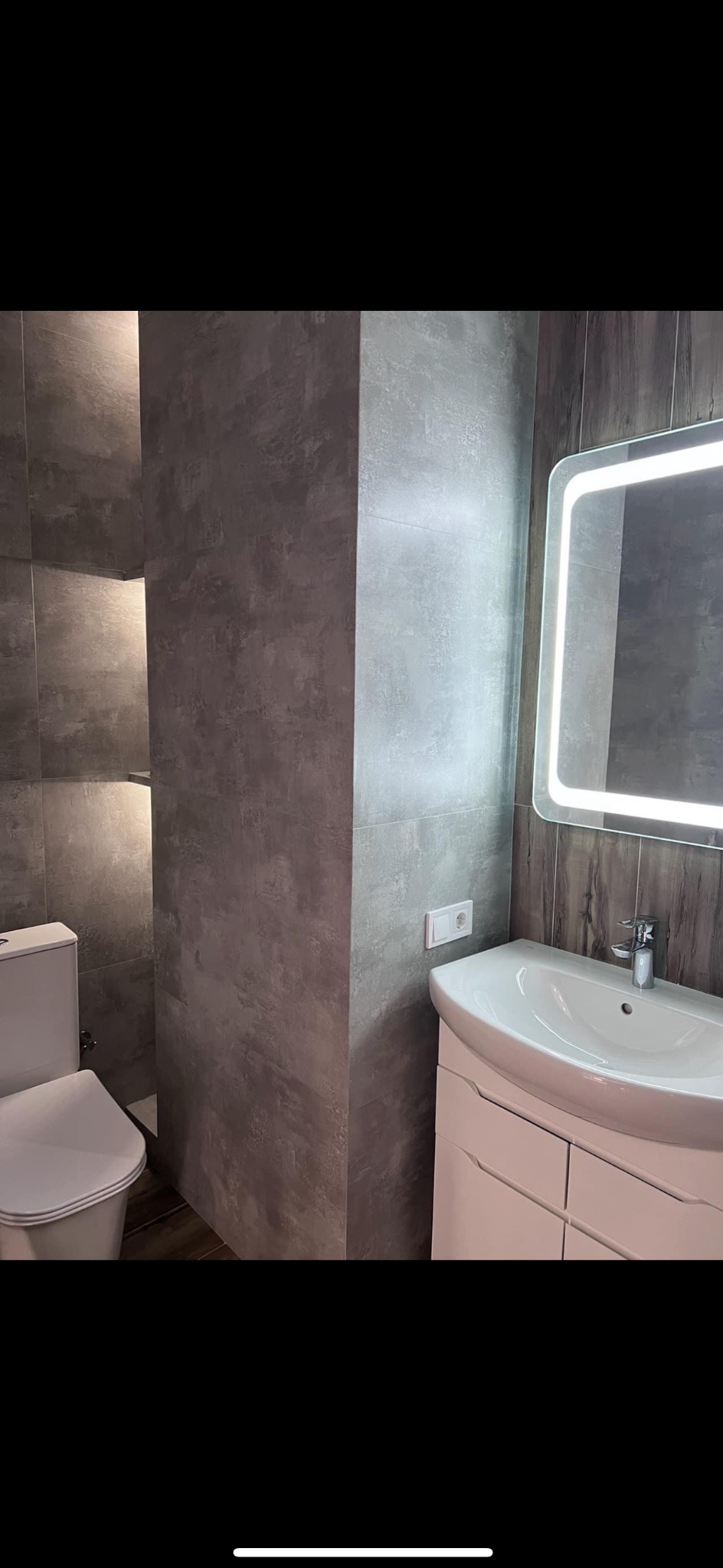 Szara łazienka z podświetlanym lustrem, widoczna toaleta i umywalka z szafką, ściany wykończone płytkami imitującymi beton.