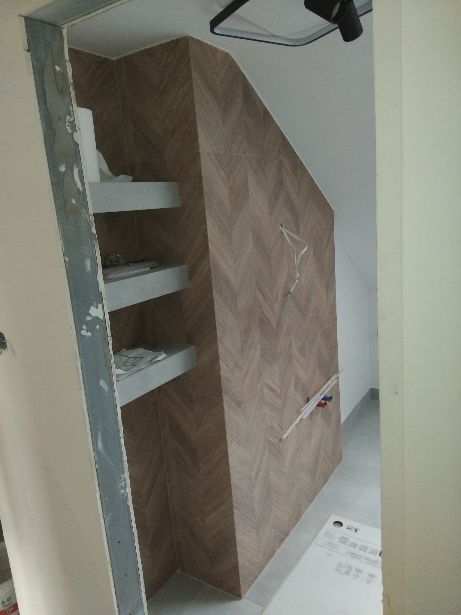 Łazienka w trakcie wykańczania: brązowe płytki ułożone we wzór jodełki na ścianie, trzy półki z szarego betonu, widoczne kable i rury instalacji wodnej, surowa ościeżnica drzwiowa.