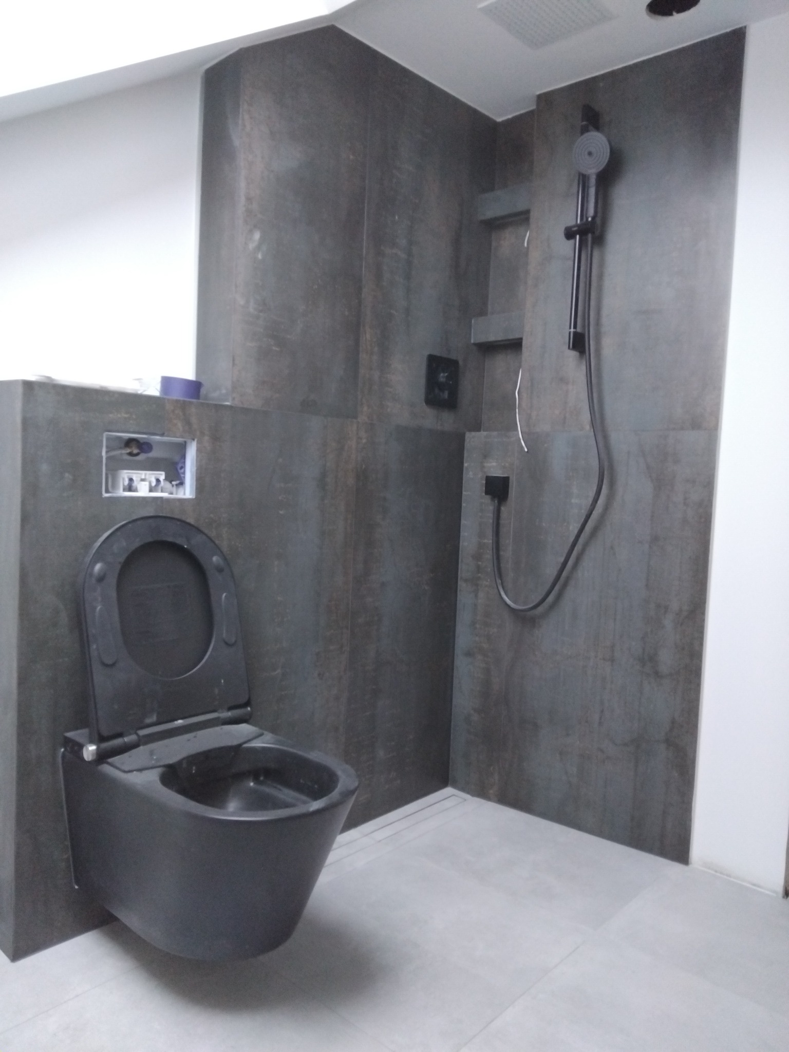 Nowoczesna łazienka z wiszącą, czarną toaletą i prysznicem typu deszczownica na ścianie wyłożonej płytkami imitującymi drewno.